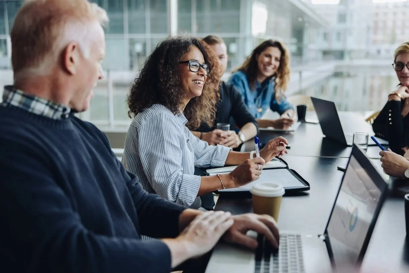 Business meeting with diverse team members sitting around a conference table, smiling and working on laptops and tablets in a modern office environment.