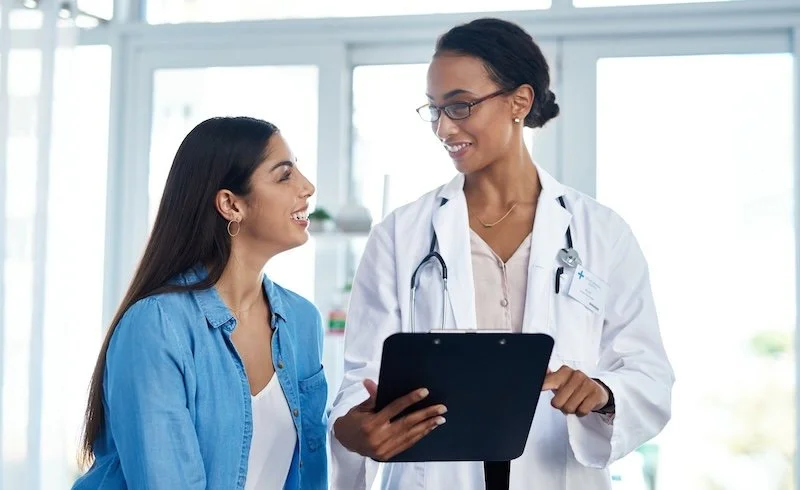 Nurse sitting in a bright clinic next to a woman patient, she is reviewing the patient's chart information with her.