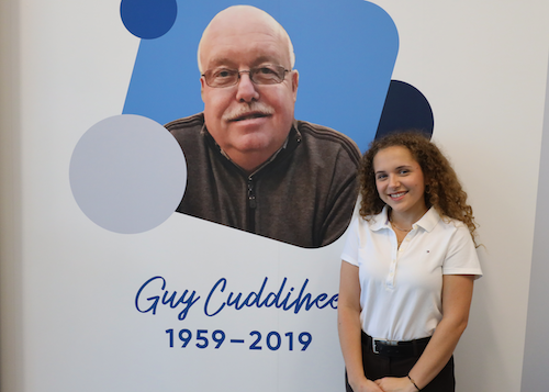 A young girl, who was the previous winner of the guy cuddihee scholarship standing in front of a memorial board with Guy Cuddihee's photo and the years 1959-2019.
