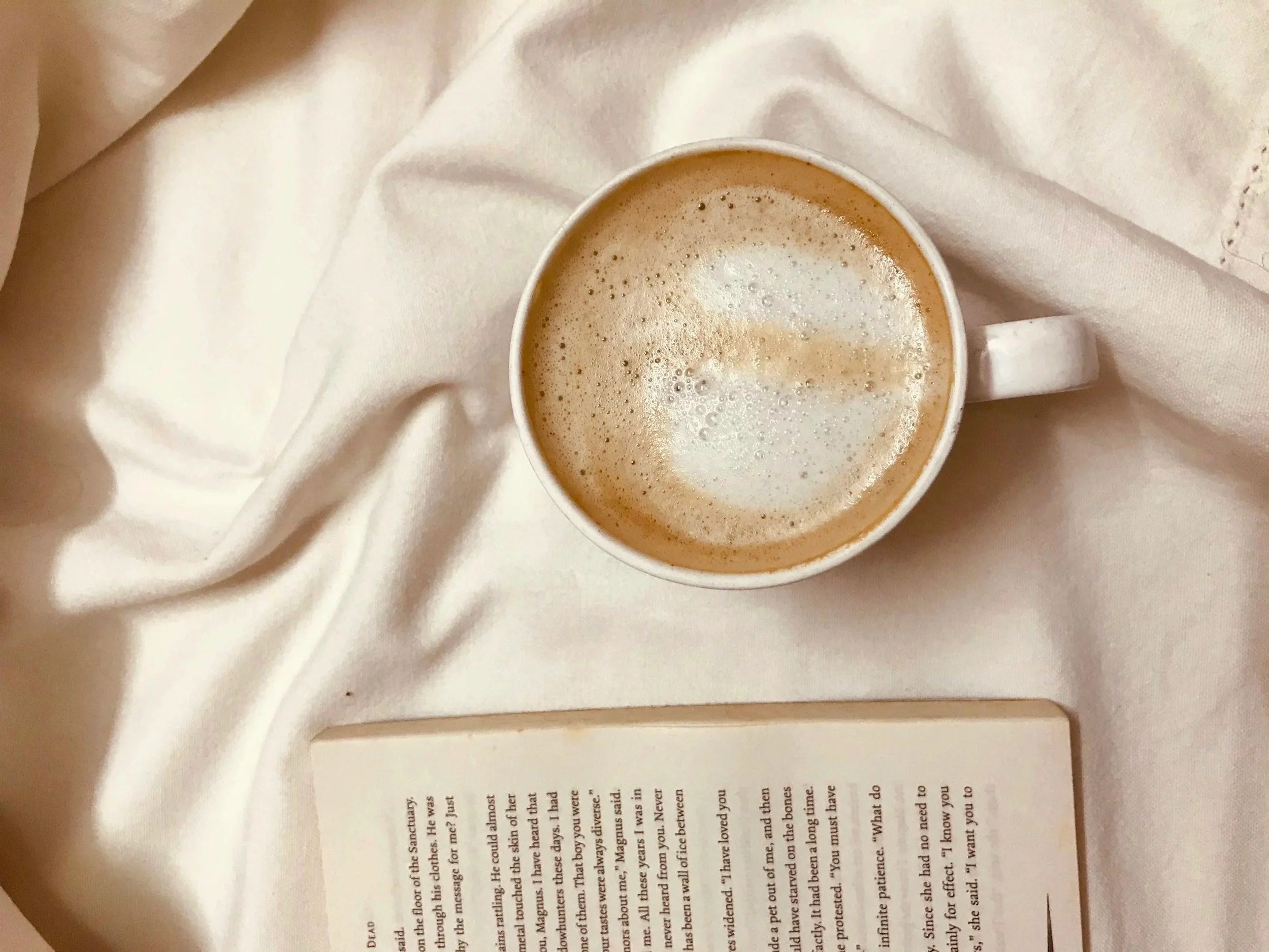 A cup of coffee with foam resting on a white fabric alongside an open book.