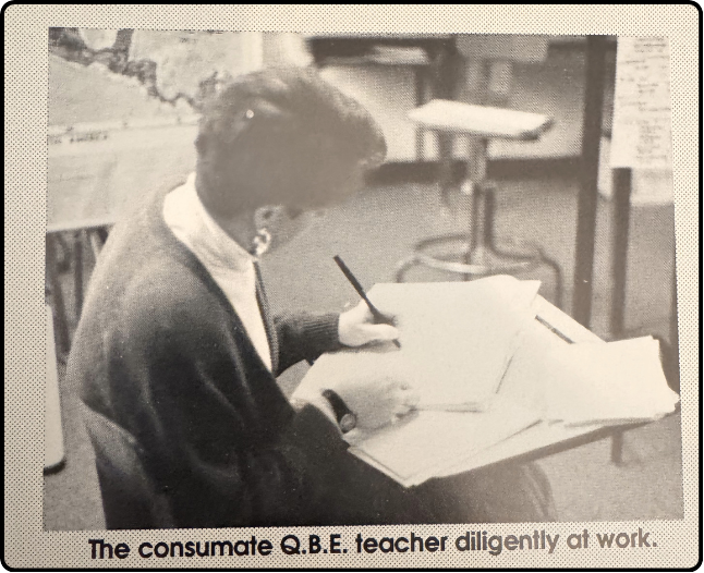 A woman with short hair, wearing a dark cardigan and light turtleneck, sitting at a desk writing with a pen in an indoor classroom setting.