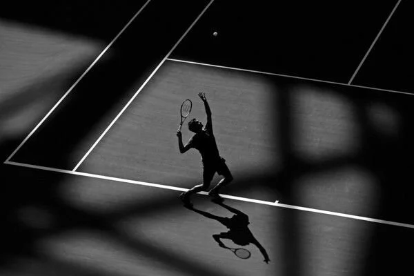 A person playing tennis on a court with a tennis ball in the air, cast in shadow with the net and court lines visible.