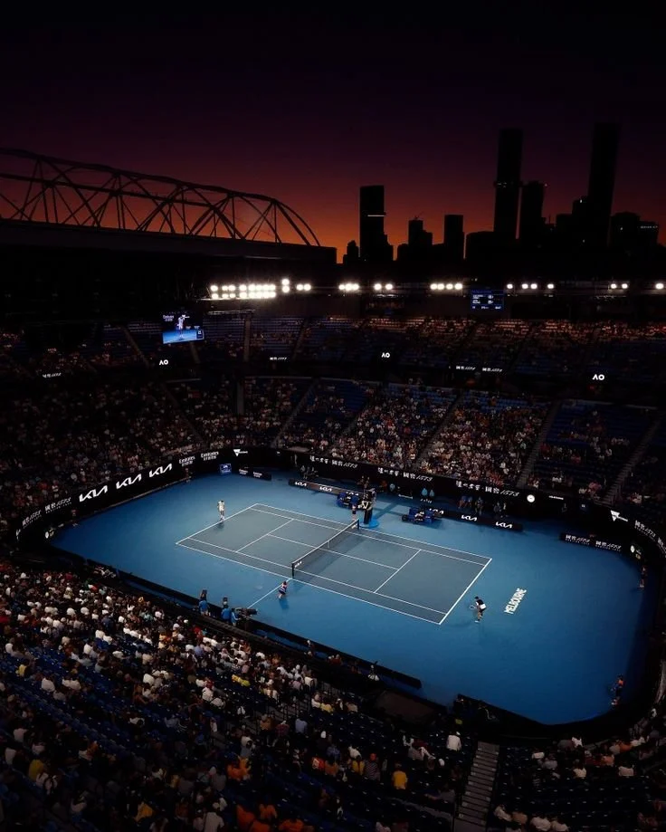 Indoor tennis court with players, audience, and city skyline at sunset in the background.