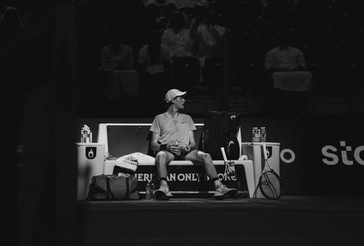 A tennis player sitting on a bench during a match, wearing a cap, T-shirt, and shorts, surrounded by tennis equipment and bottles, with an audience in the background.
