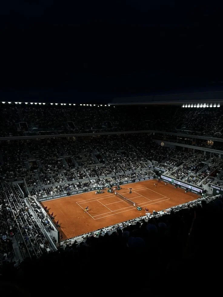 An indoor clay tennis court with players and a small audience in a dimly lit arena.