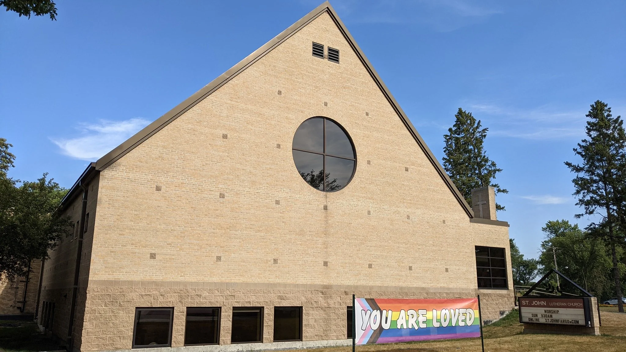 a photo of the outside of the St. John Lutheran Church building. There is a banner tied between stakes on the lawn with the text You Are Loved on a background in the colors of the progress pride flag.