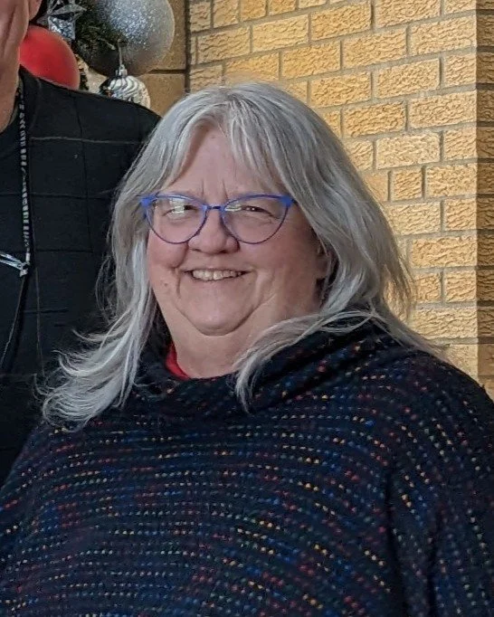 a woman with long grey hair and glasses smiling at the camera.