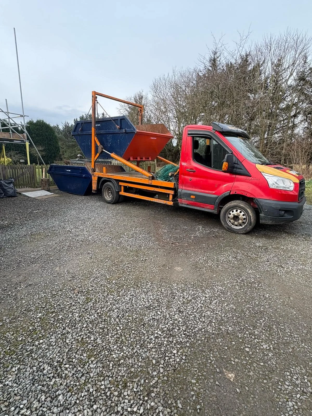 A small red flatbed truck parked outdoors with two large blue and red dumpsters on its bed, surrounded by gravel and trees in the background.