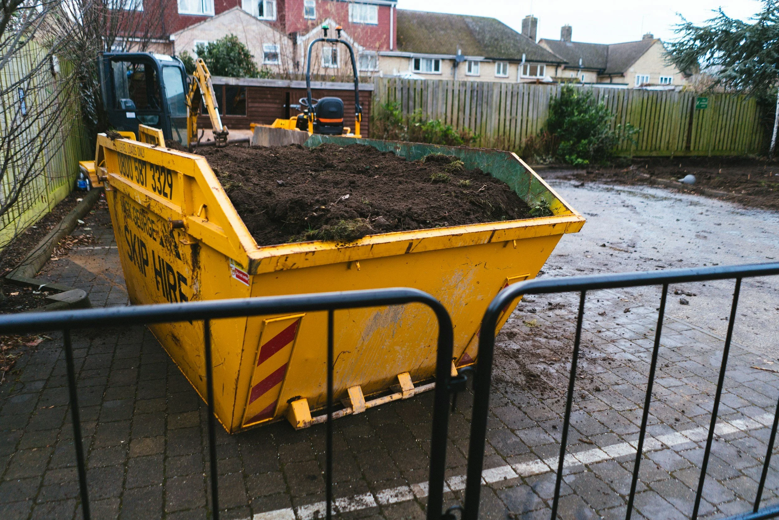 A yellow construction skip filled with dirt placed on a paved sidewalk at a construction site surrounded by a metal fence, with some small excavator machinery and houses in the background.
