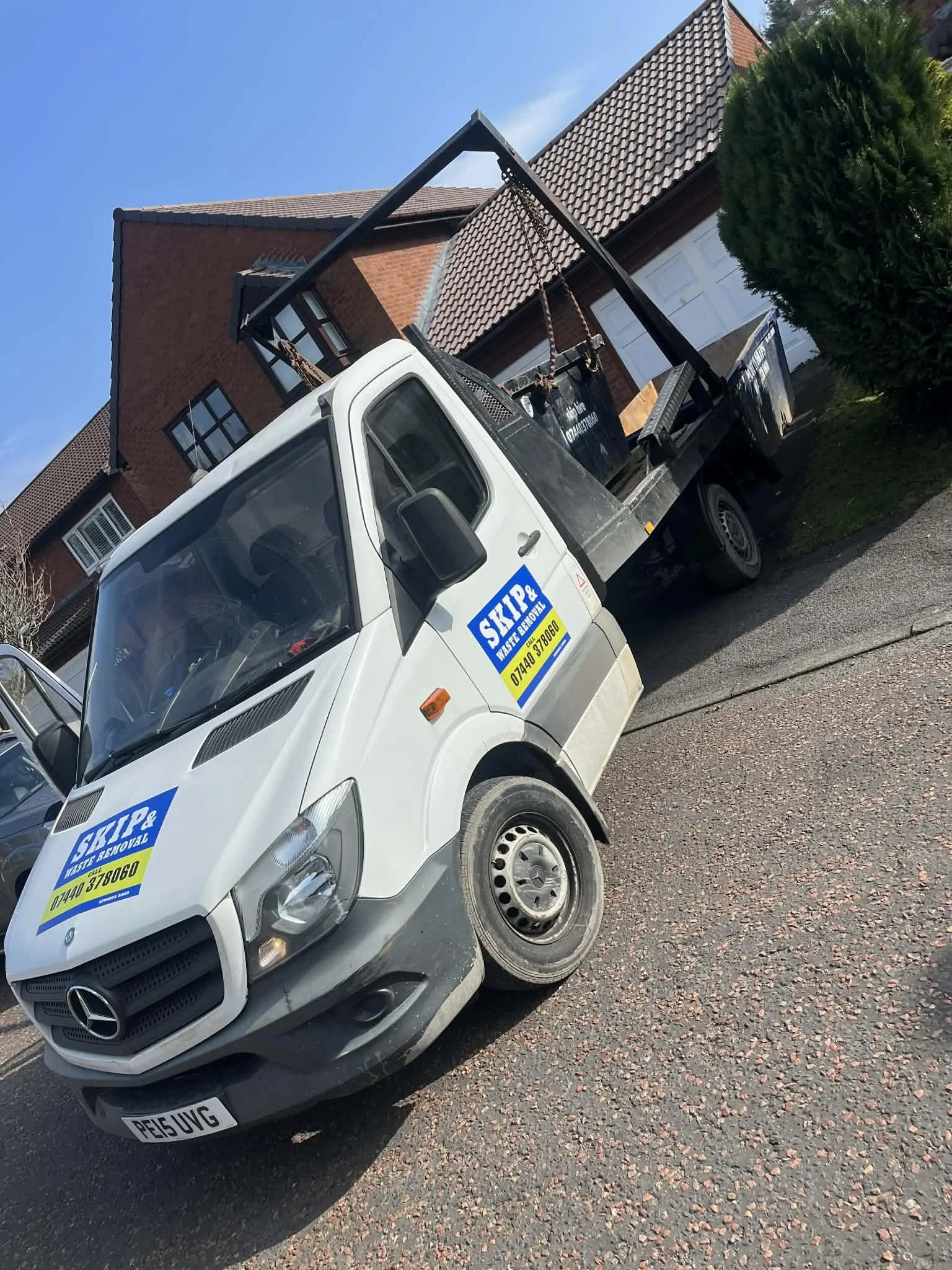 A small white tow truck parked on a residential street with houses in the background. The truck has signage that reads "Skip & Waste Removal" and a phone number. The truck is positioned at an angle on the pavement.