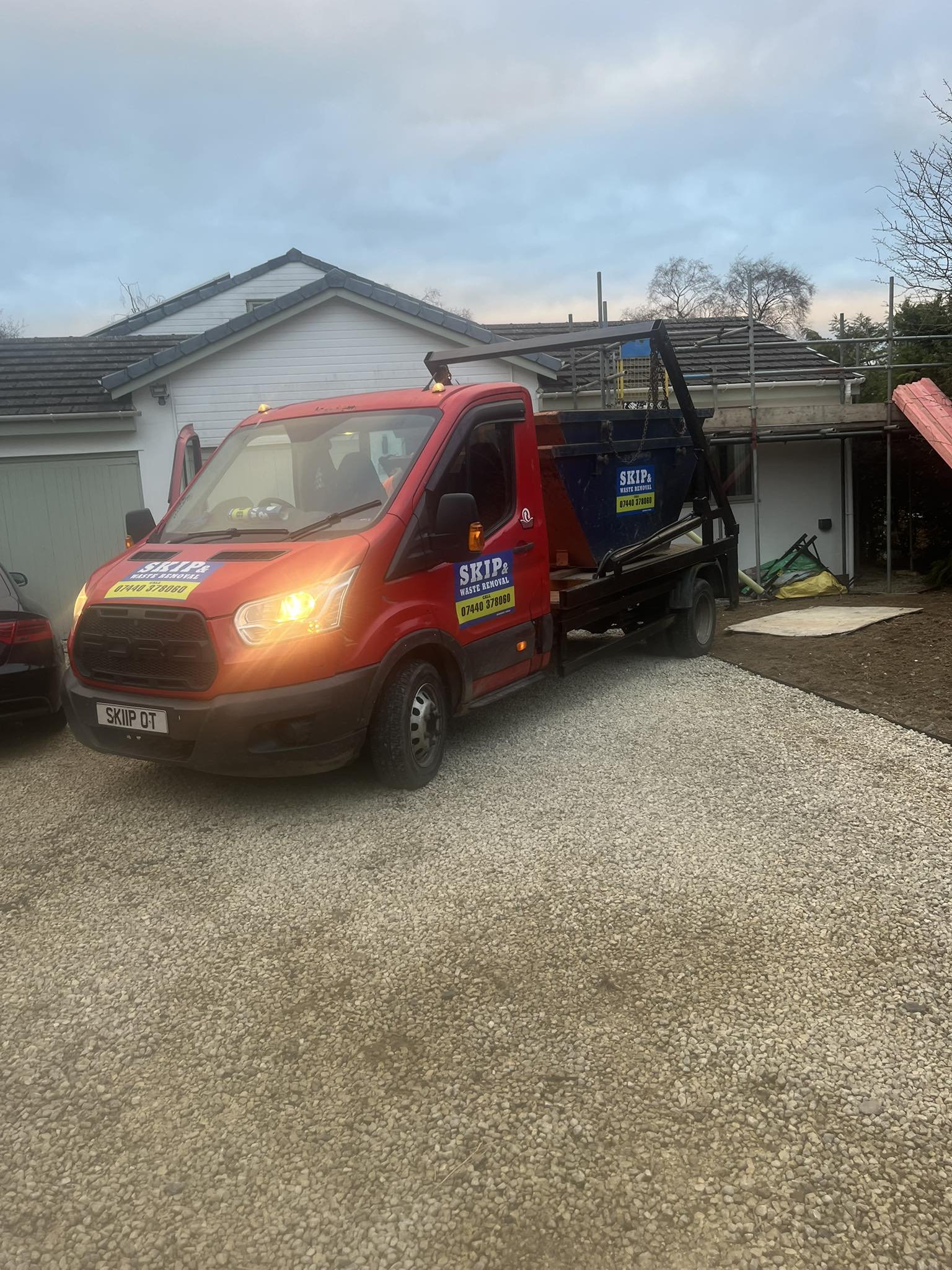 A red waste removal truck with company logos and contact information parked on a gravel driveway in front of a house under construction or renovation.