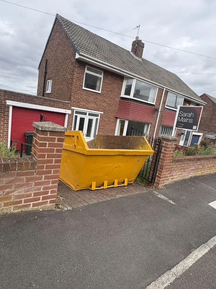 Large yellow dumpster placed on the sidewalk outside a brick house with a sign that reads "Sarah Mains."