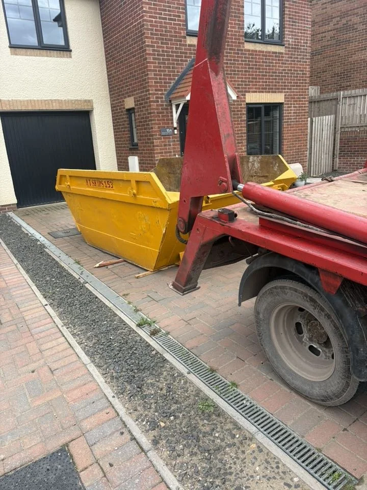A yellow construction dumpster attached to a red tow truck parked beside a brick residential house.