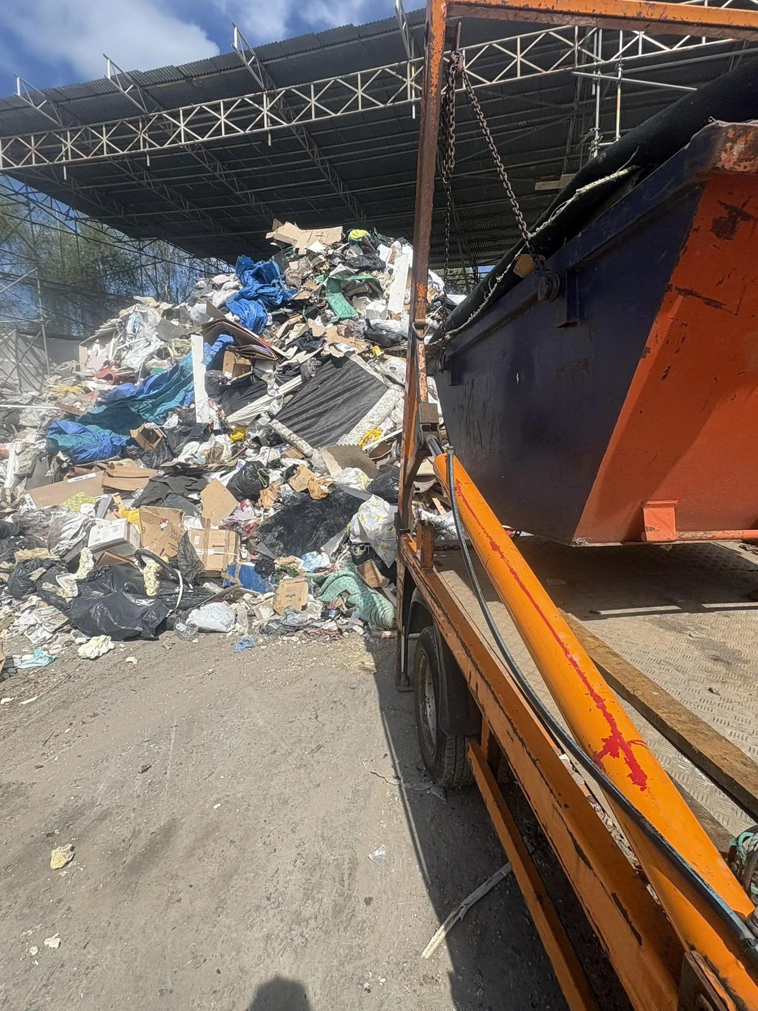 A large pile of mixed trash and debris, including cardboard, plastic bags, and miscellaneous waste, is being loaded or transported by an orange waste management truck at an outdoor recycling or waste disposal facility.