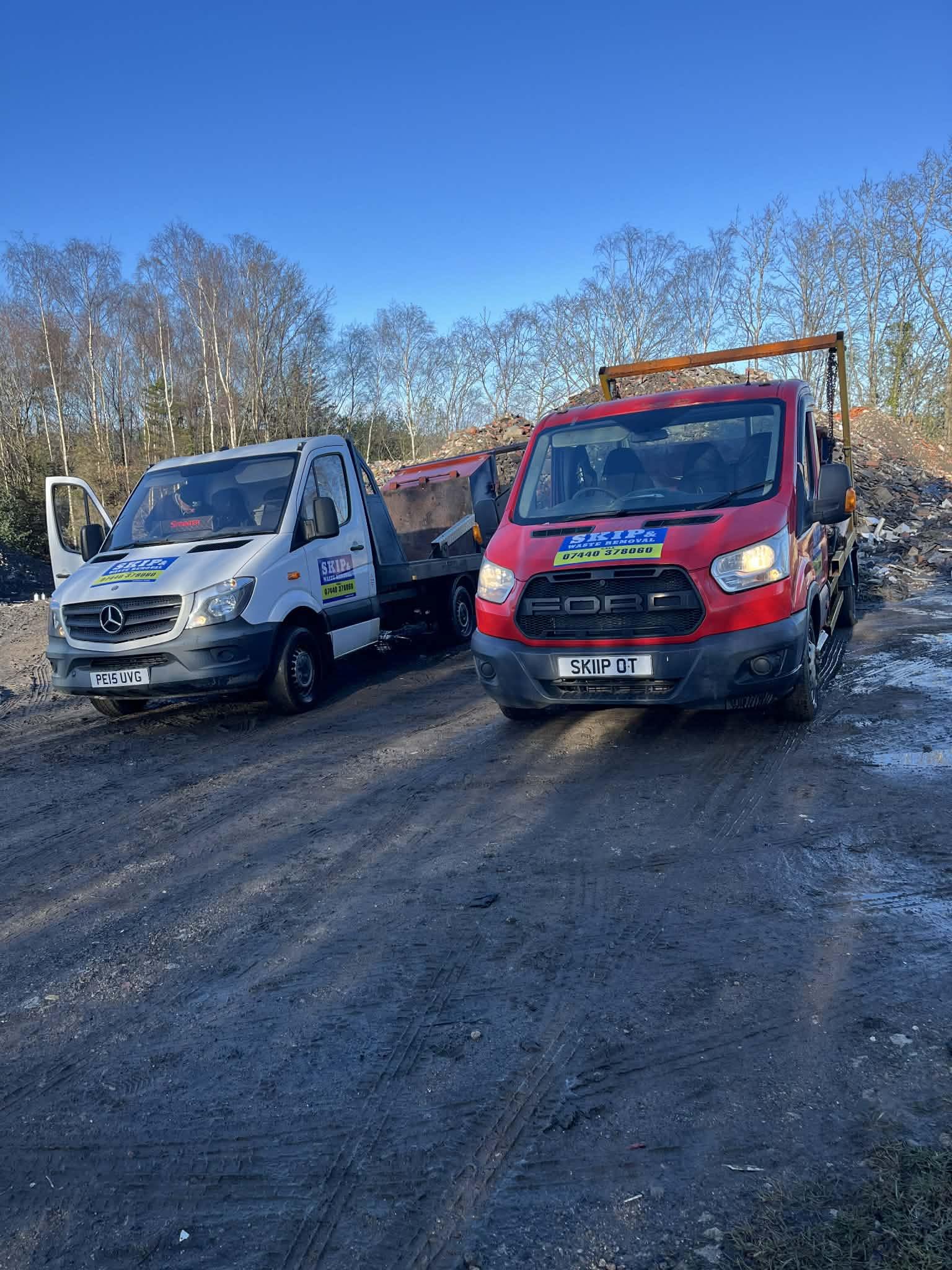 Two trucks parked on a dirt surface with a backdrop of leafless trees under a clear blue sky.