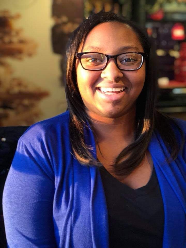 A woman with black hair and glasses smiling at the camera, wearing a blue jacket and a black shirt, in an indoor setting.