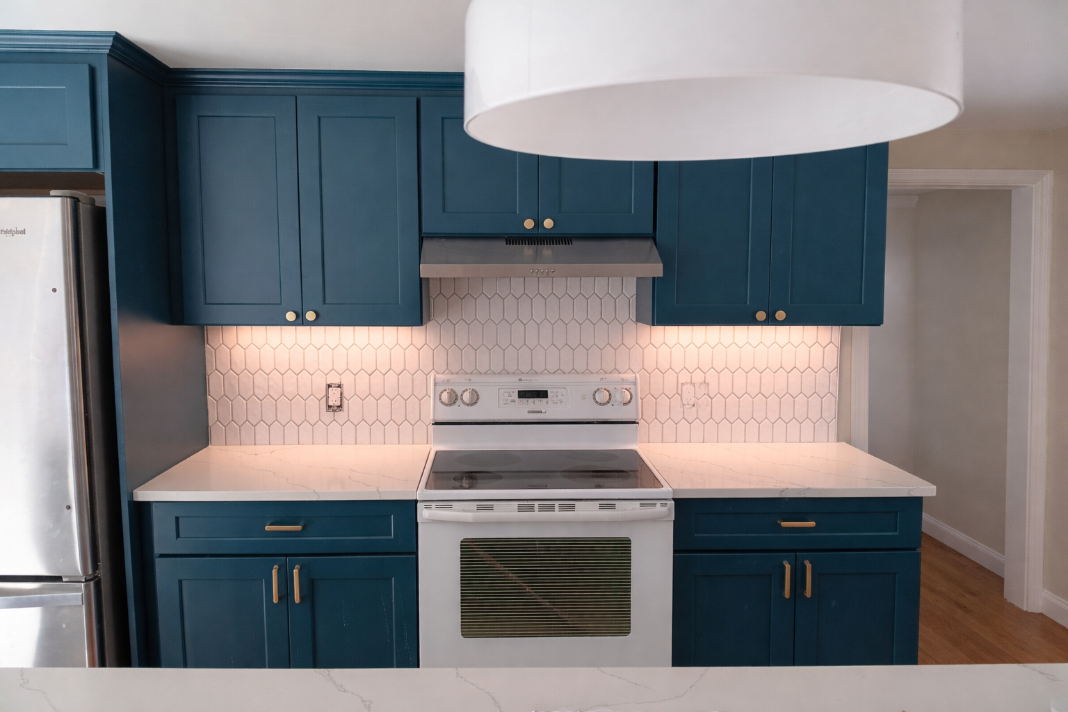 Kitchen with blue cabinets, white marble countertop, hexagonal tile backsplash, white stove, stainless steel refrigerator on the left, and a large white pendant light overhead.