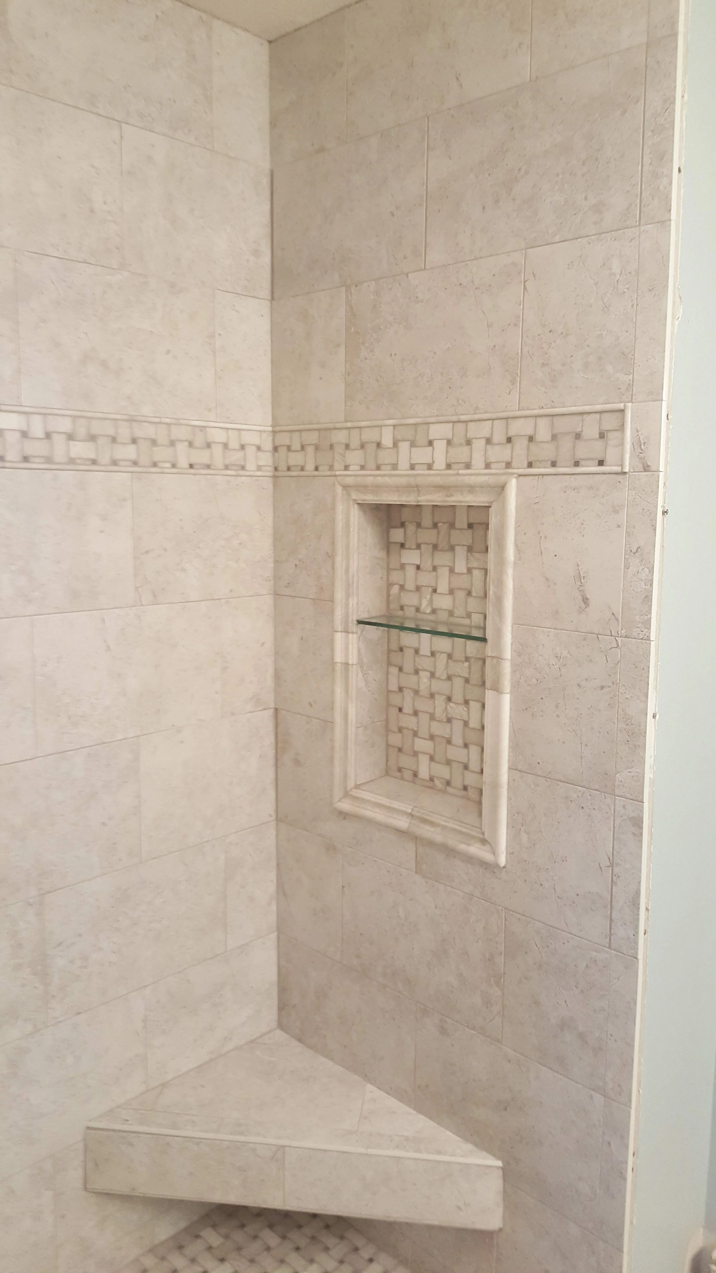 A corner of a tiled shower with a built-in recessed shelf with a glass shelf and decorative tile accents.