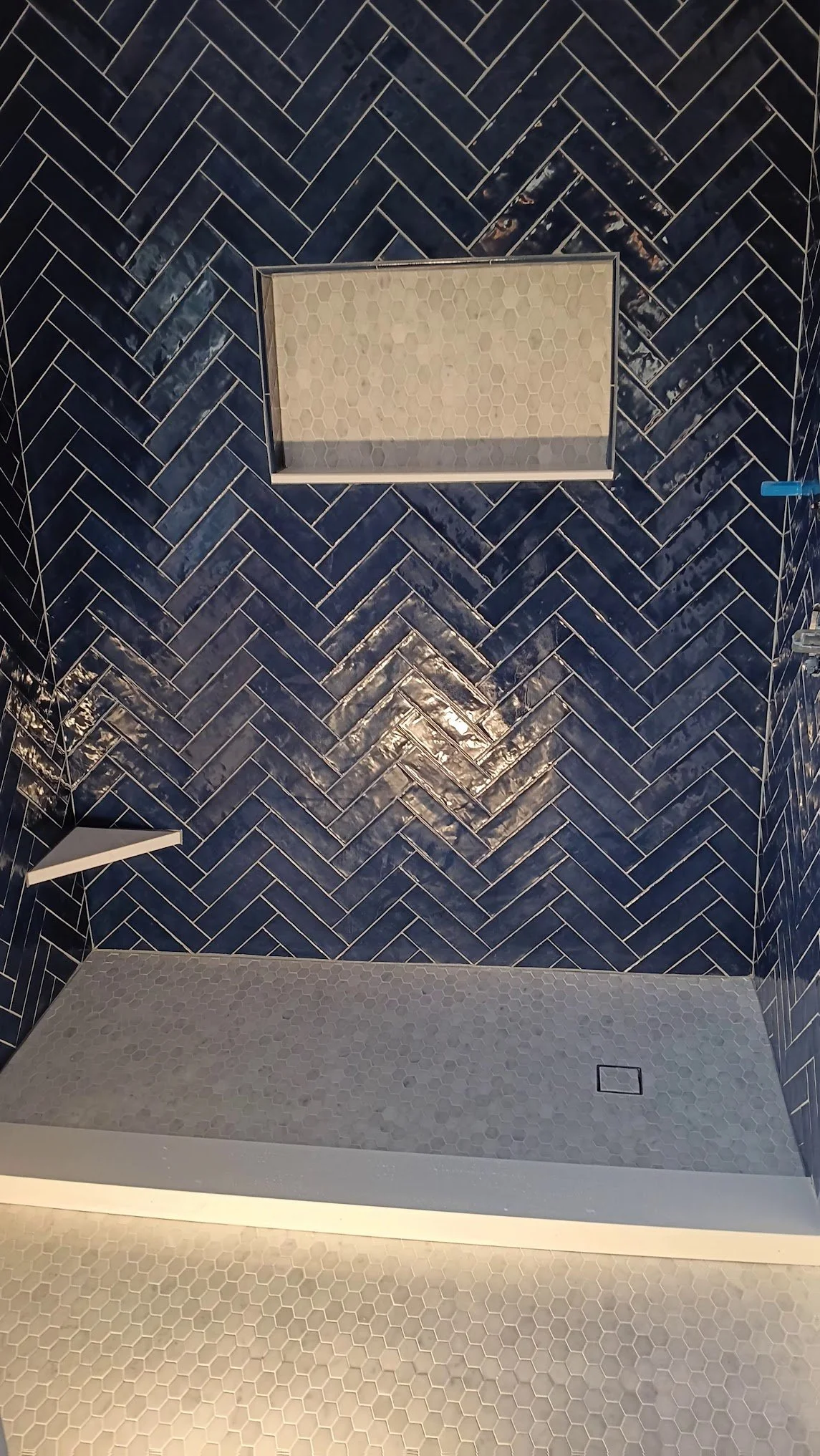 Shower with dark blue herringbone tiled wall, white hexagon tiled floor, and a small recessed shelf with white hexagon tiles.