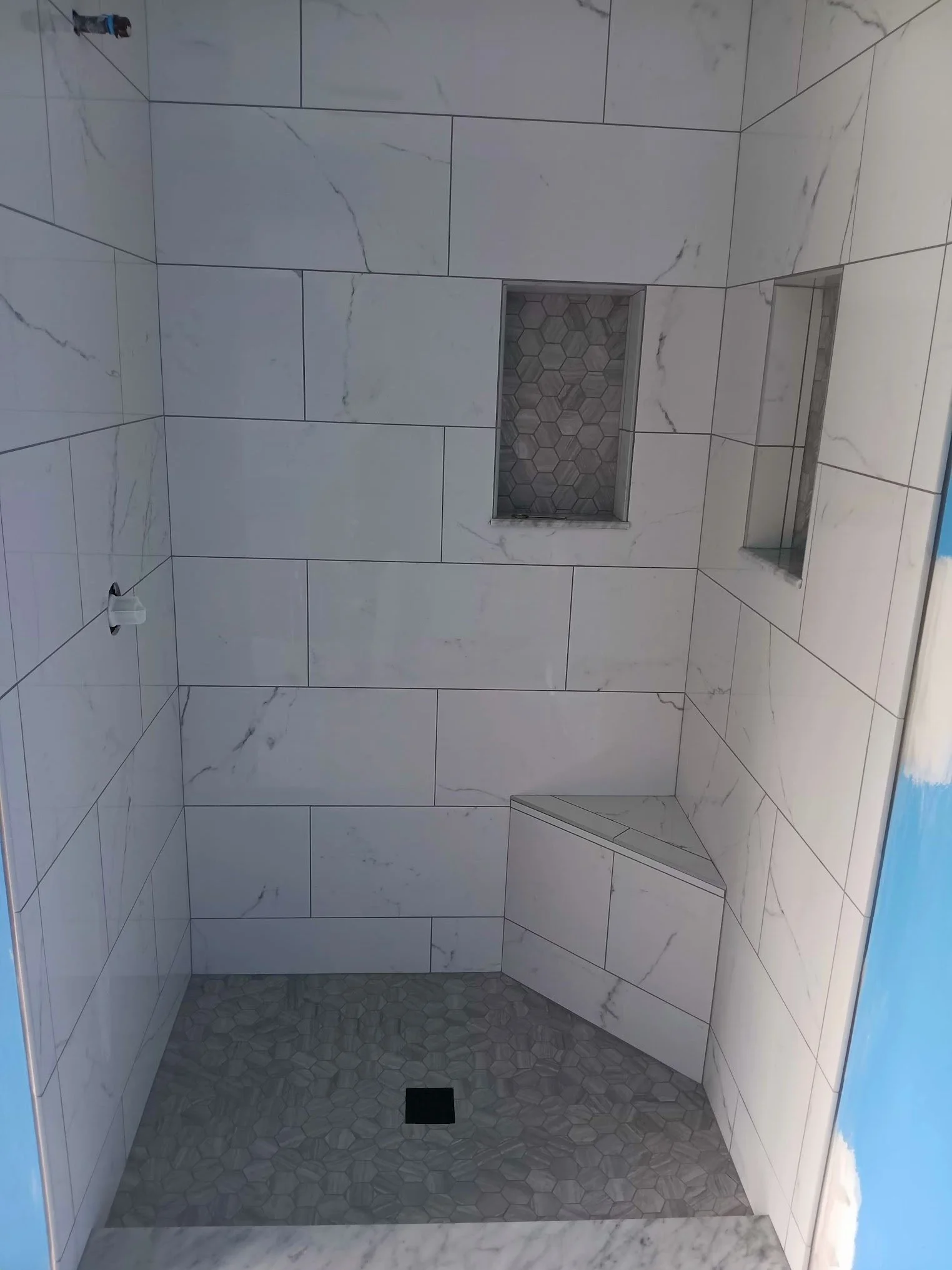 View of a tiled shower with large white marble tiles on the walls, hexagonal small tiles on the shower floor, a built-in niche with hexagonal tiles, and a black drain at the floor's center.
