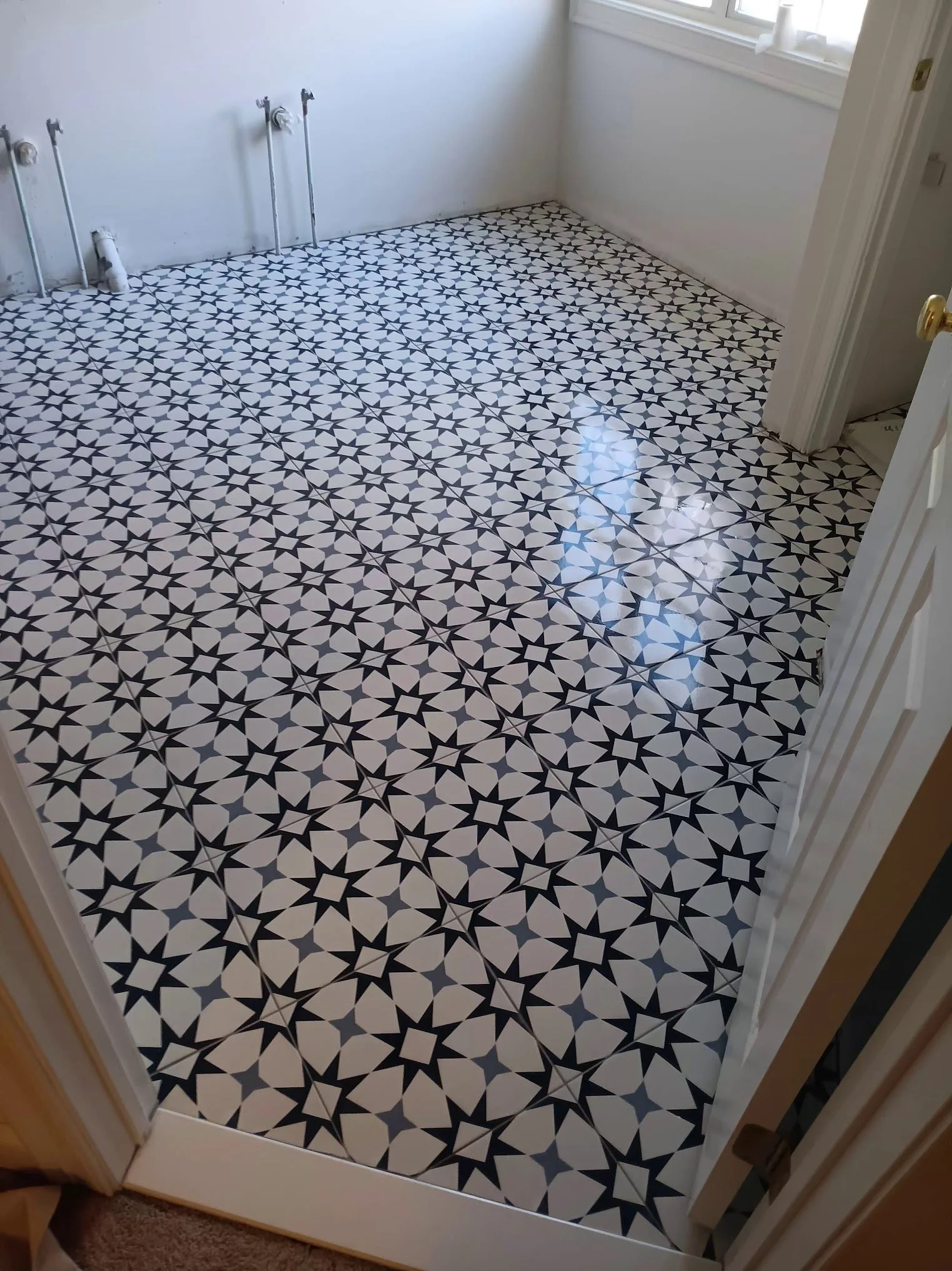 Room with black and white patterned tile floor, white walls, a window with white trim, and plumbing fixtures along the wall, indicating a space under renovation or construction.