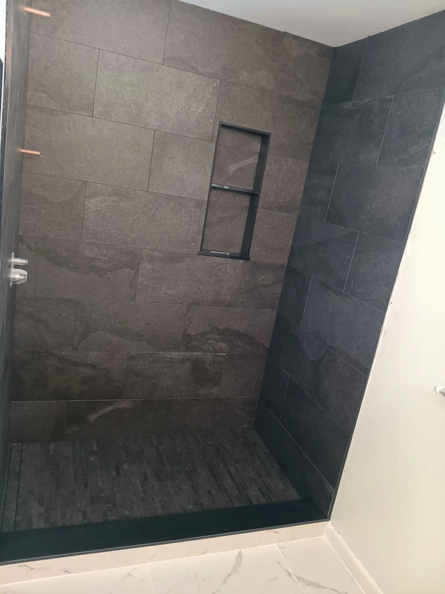 A modern bathroom shower area with dark gray stone tiles, a built-in niche for toiletries, and a glass door. The wall and floor tiles are different shades of gray, with the floor tiles having a denser pattern. The edge of the shower is lined with whi