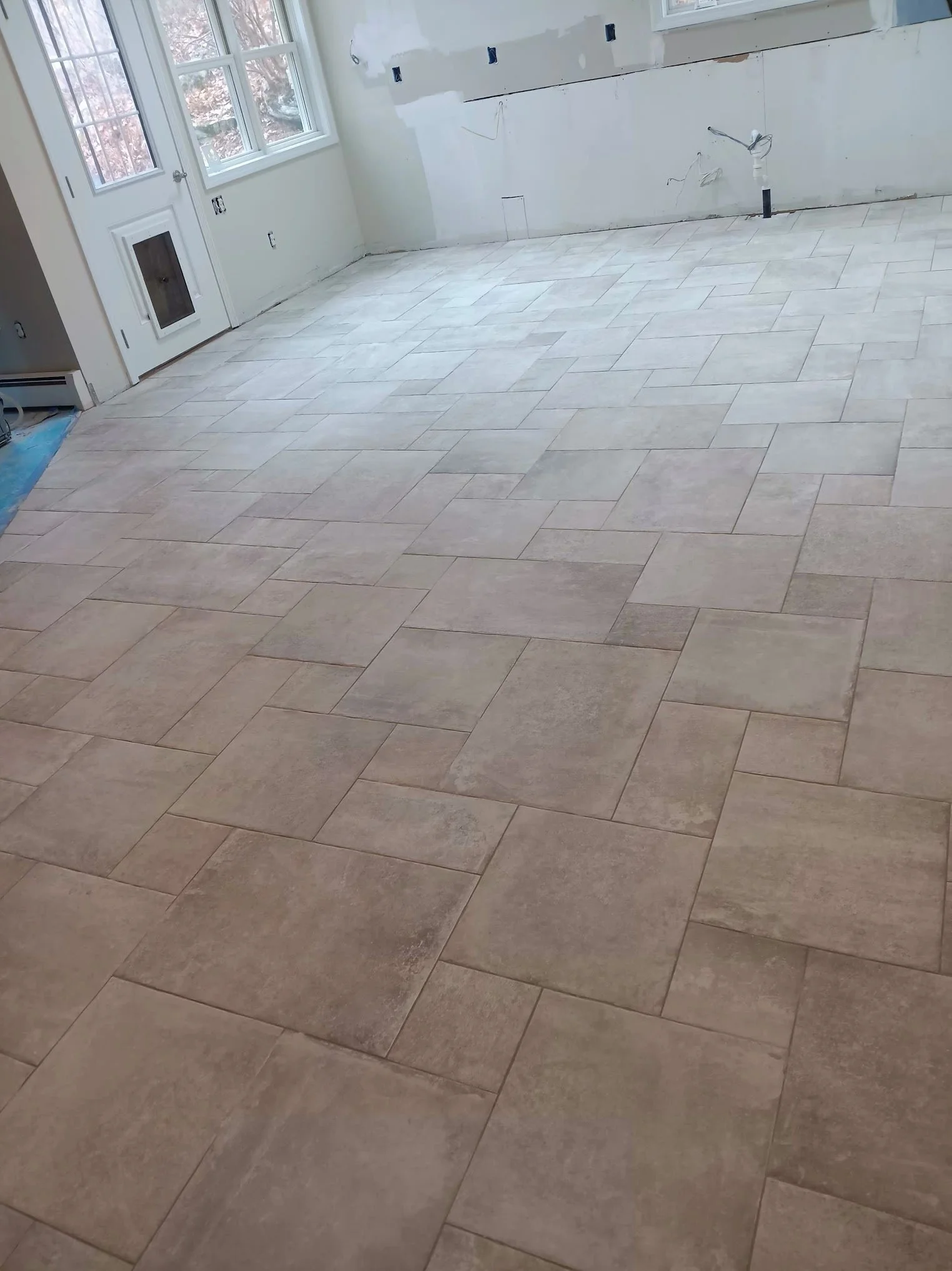 A room under renovation with beige tile flooring, unfinished white walls, and a window showing an outdoor scene of trees. There are electrical wires and outlets visible on the walls.
