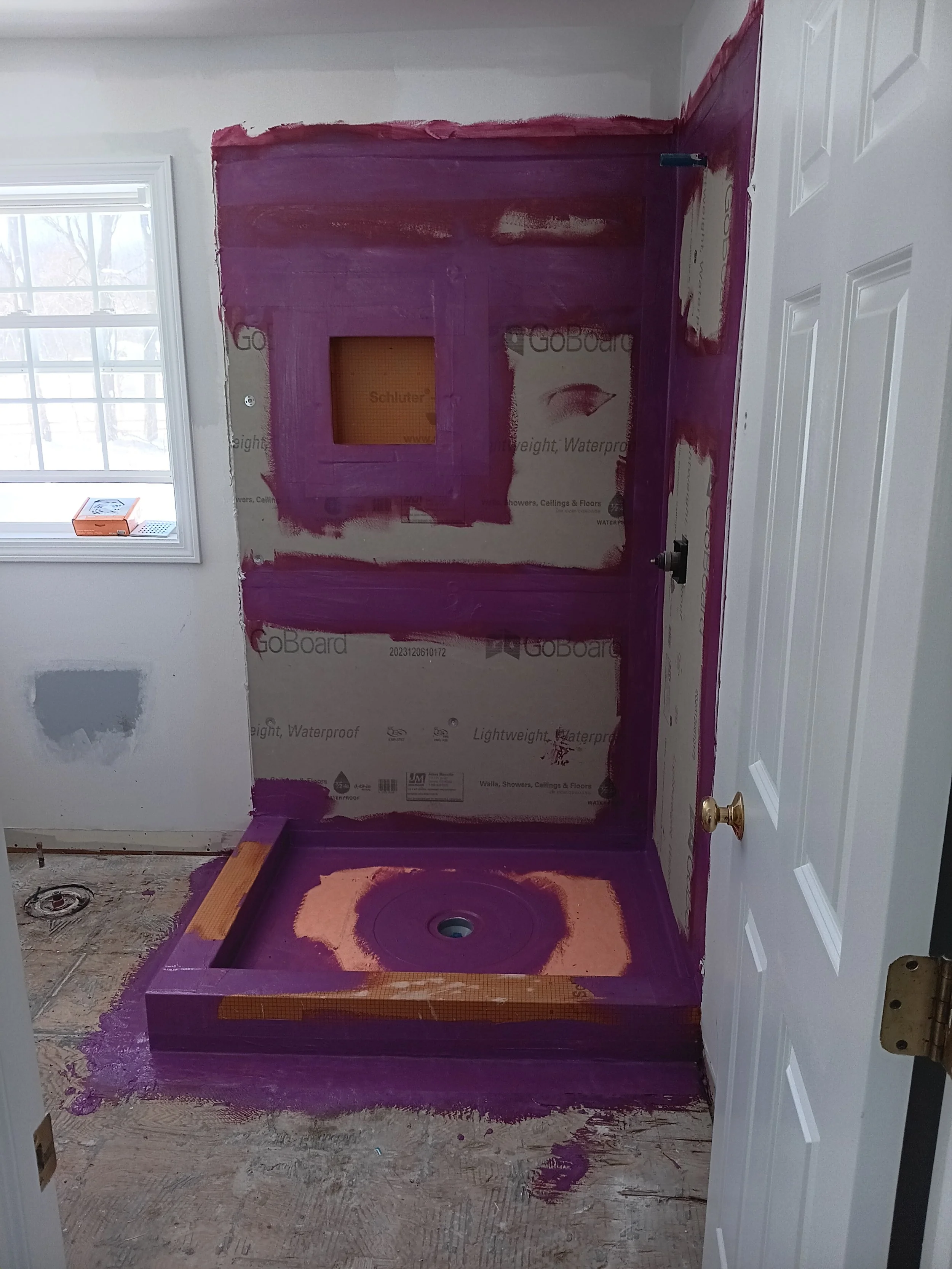 Bathroom under renovation with purple waterproofing paint applied around the shower area, and the floor stripped down to the subfloor.