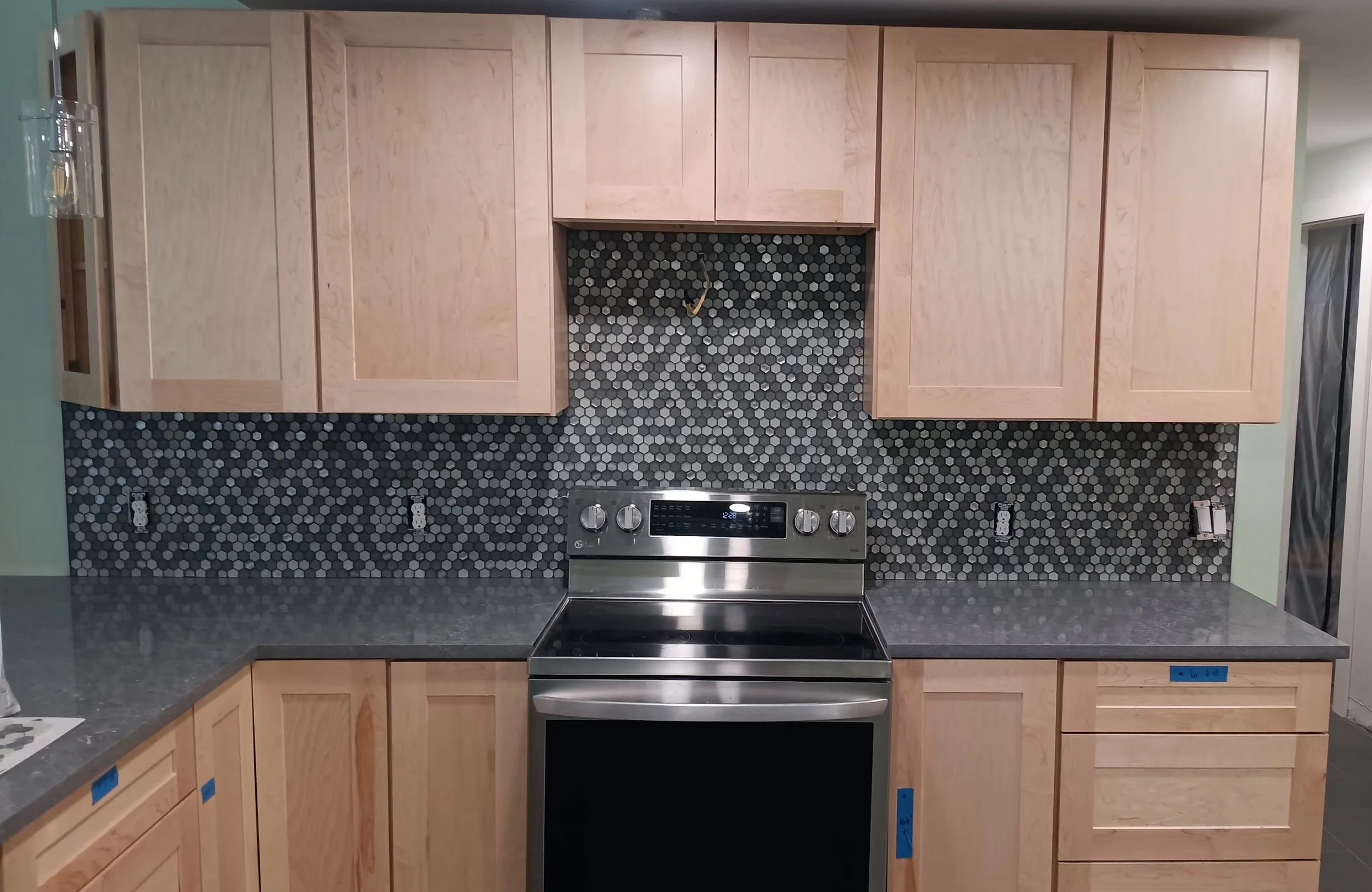A kitchen display with light wooden cabinets, a black and gray honeycomb backsplash, and a stainless steel electric stove with oven. The countertop is gray, and there is a small amount of blue painter's tape on the lower cabinets.