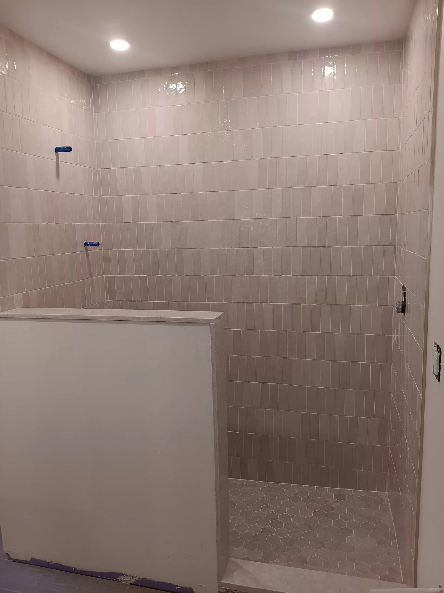 A bathroom shower area with light pink rectangular tiles on the walls, a white partition wall at the entrance, and hexagonal tiles on the floor, with two recessed ceiling lights overhead.