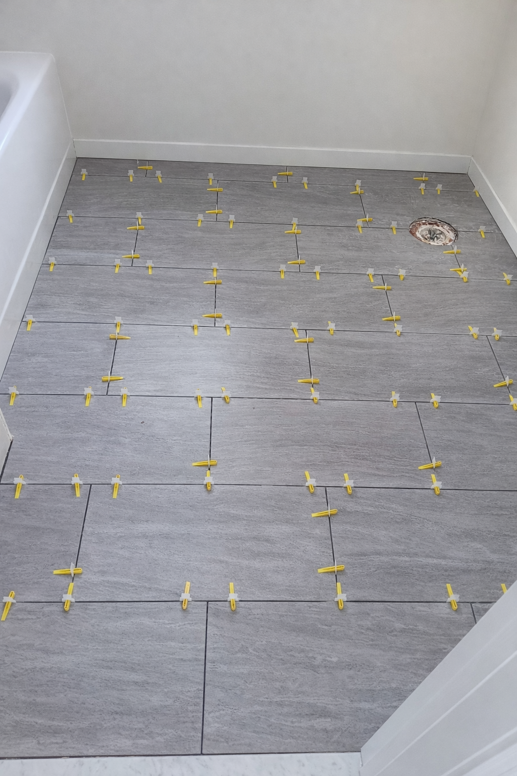 Bathroom floor with gray tiles, yellow tile spacers in place, and a drain hole in the upper right corner.