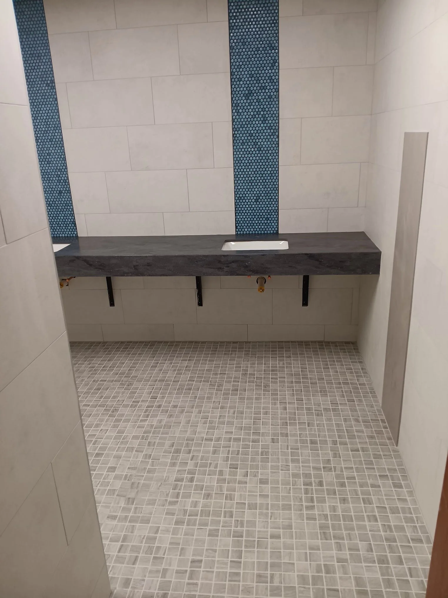 A bathroom with light-colored wall tiles, a dark gray stone countertop, and a tiled floor. The wall tiles include decorative blue mosaic stripes. There is a sink cutout in the countertop, and plumbing fixtures are visible beneath it.
