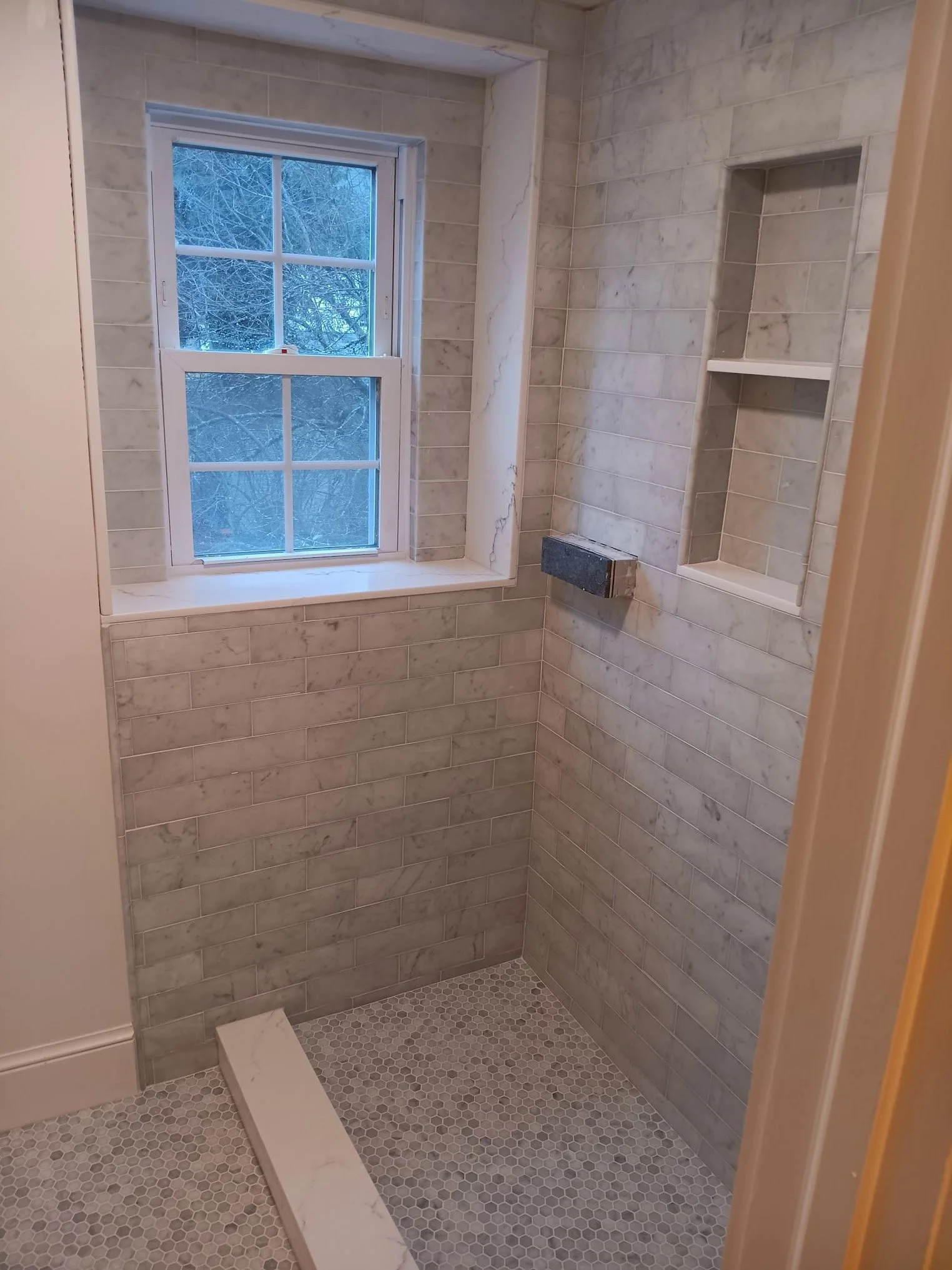 A bathroom shower area with light-colored marble tile walls, a small window with a winter scene outside, a built-in shelf, a recessed vent or niche, and a gray hexagon tile floor.