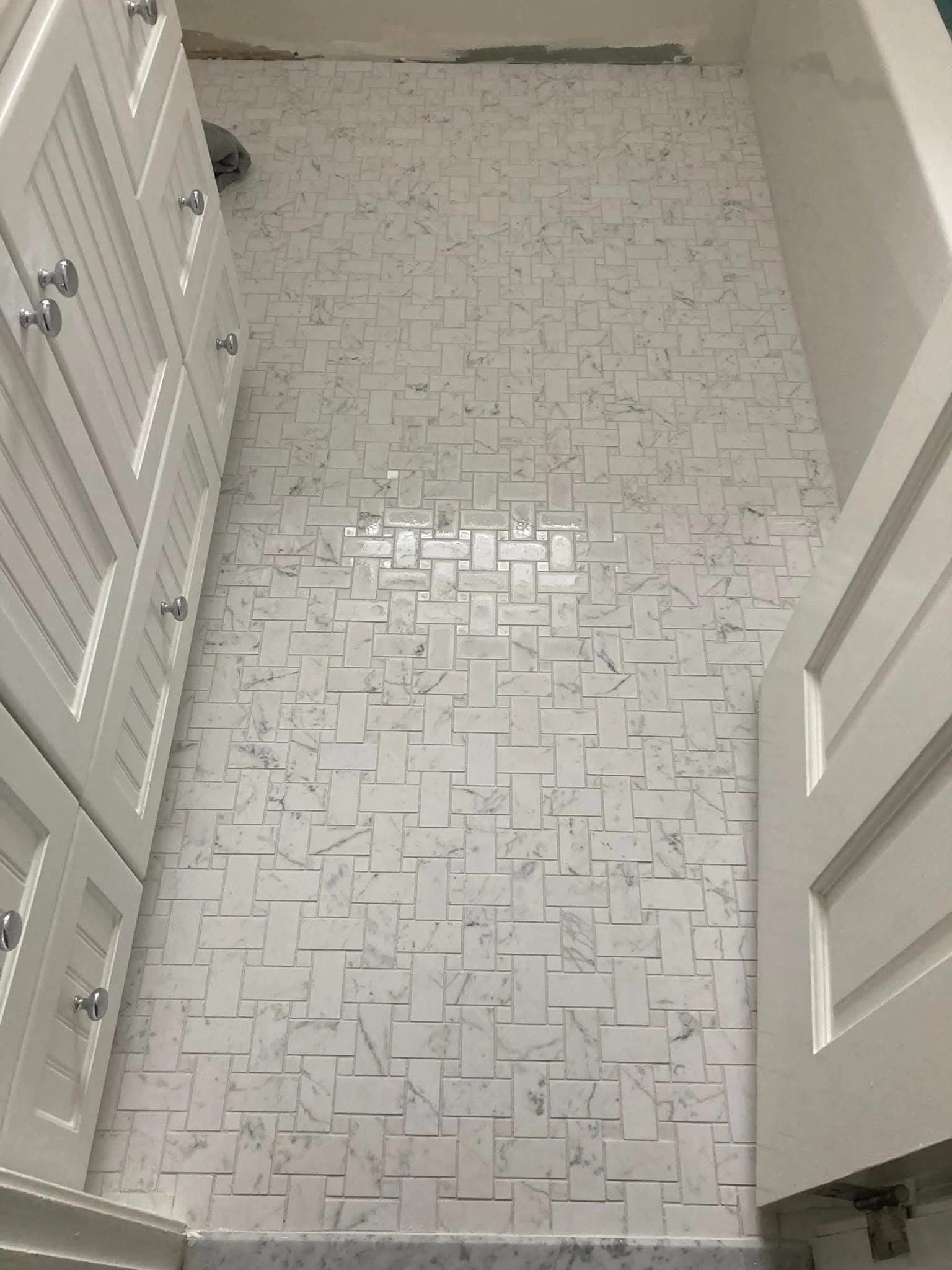 Close-up photo of a bathroom floor with white marble tiles in a herringbone pattern, with a white cabinet on the left and a curved white wall on the right. The space appears to be under renovation.