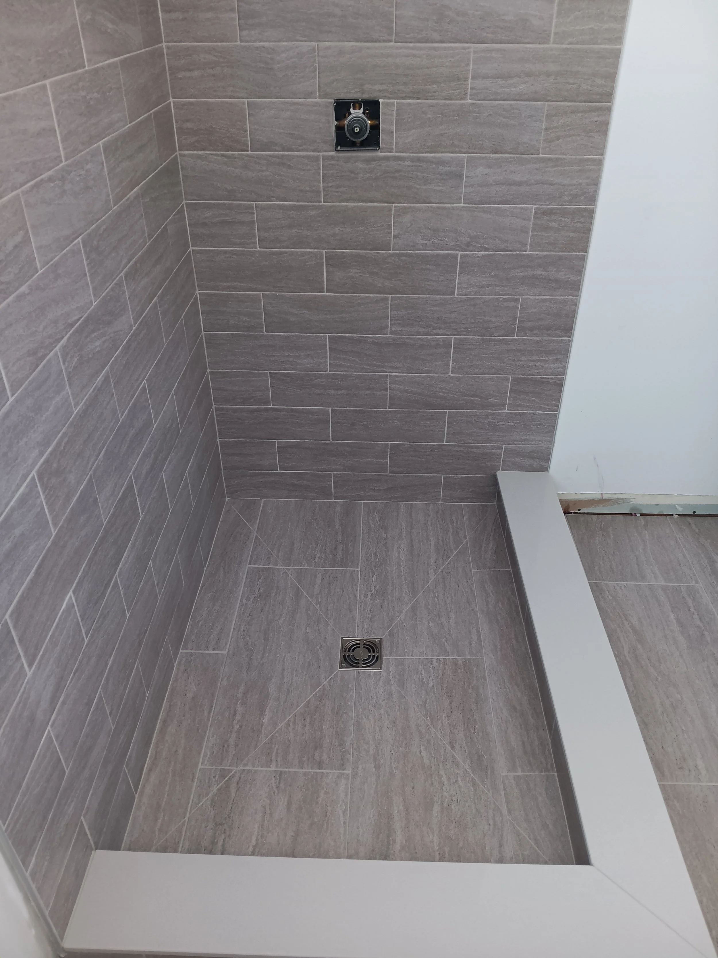 Empty shower stall with gray tiles, a drain on the floor, and an unfinished fixture hole in the wall.