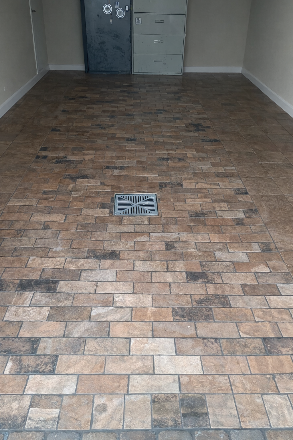 Indoor room with brick-patterned tile flooring, a metal drain in the center, beige walls, and a gray metal door with a lock and two circular windows, along with a gray set of drawers in the background.