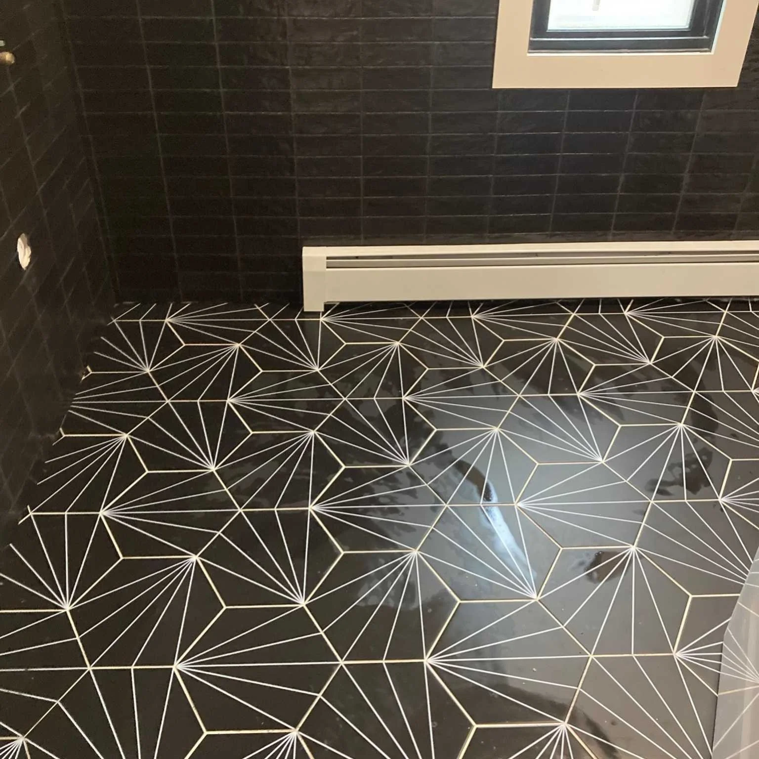 Close-up of a tiled floor with a geometric pattern of white lines on black tiles, next to a dark tiled wall and a baseboard heater below a window.