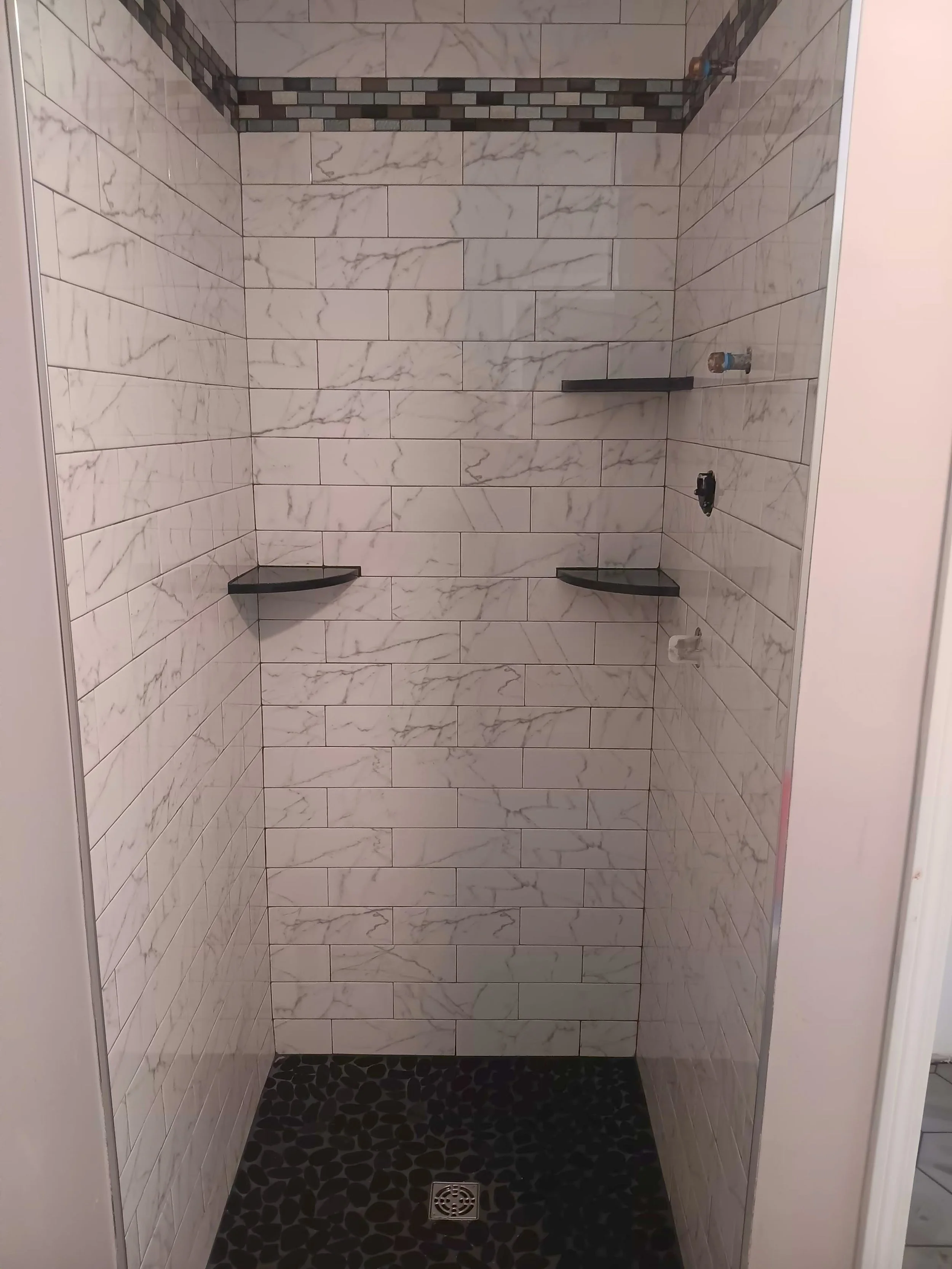 Empty shower with white marble tiles on the walls, black pebble floor, and a small black shelf inside.
