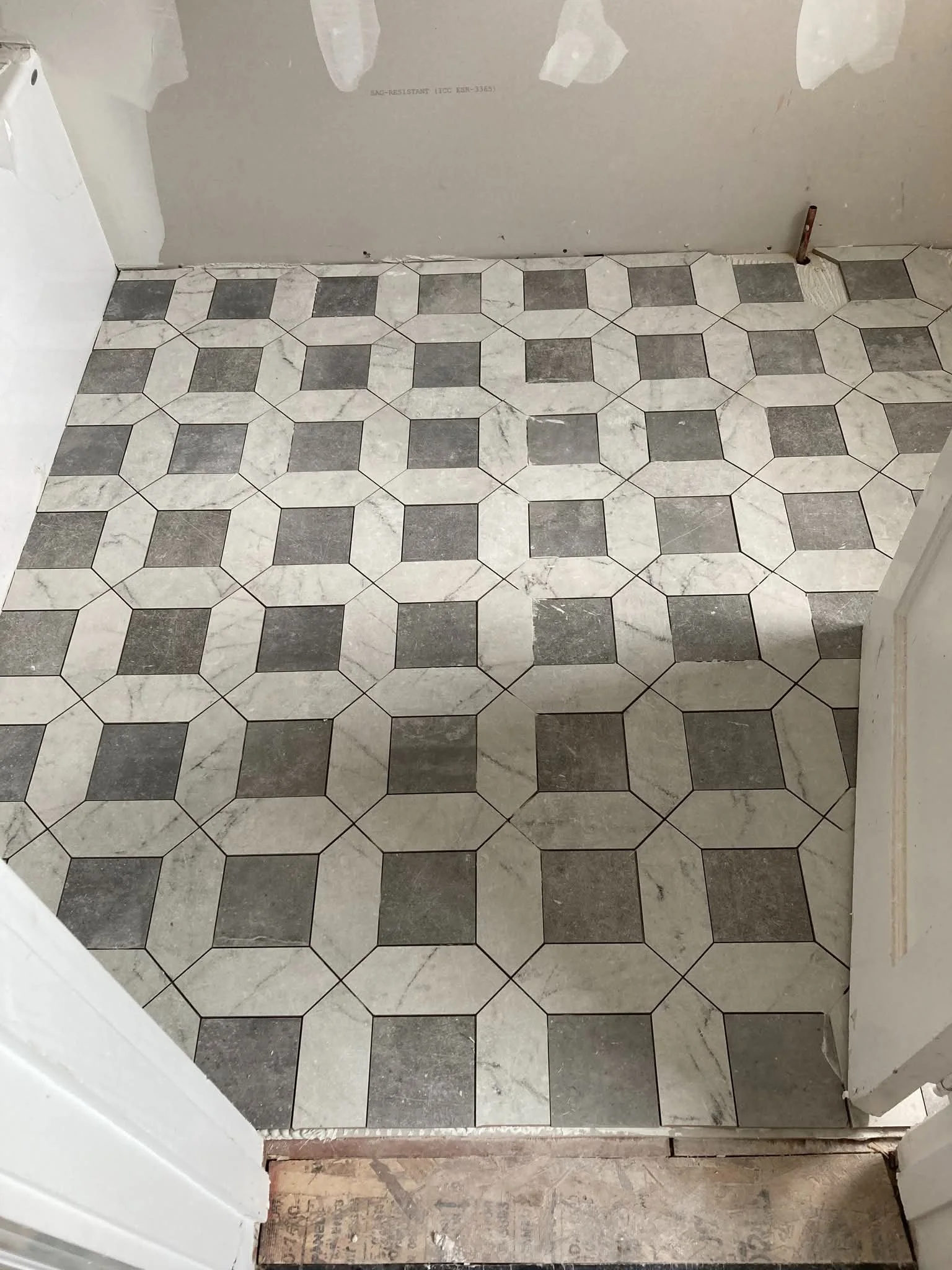 A partially renovated room with tile flooring featuring a repetitive pattern of octagons and squares in shades of gray and white. The walls are unfinished, with visible drywall and joint compound.