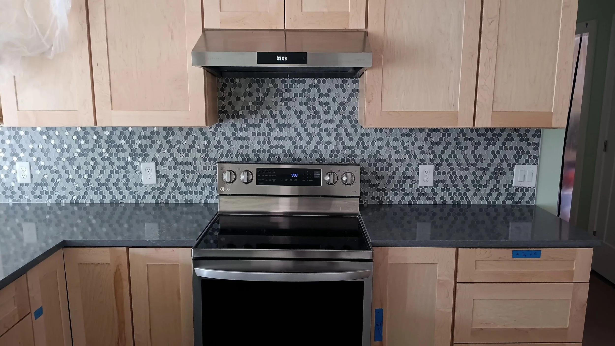 Kitchen with light wood cabinets, grey hexagon tile backsplash, black countertop, stainless steel LG oven, and white electrical outlets.