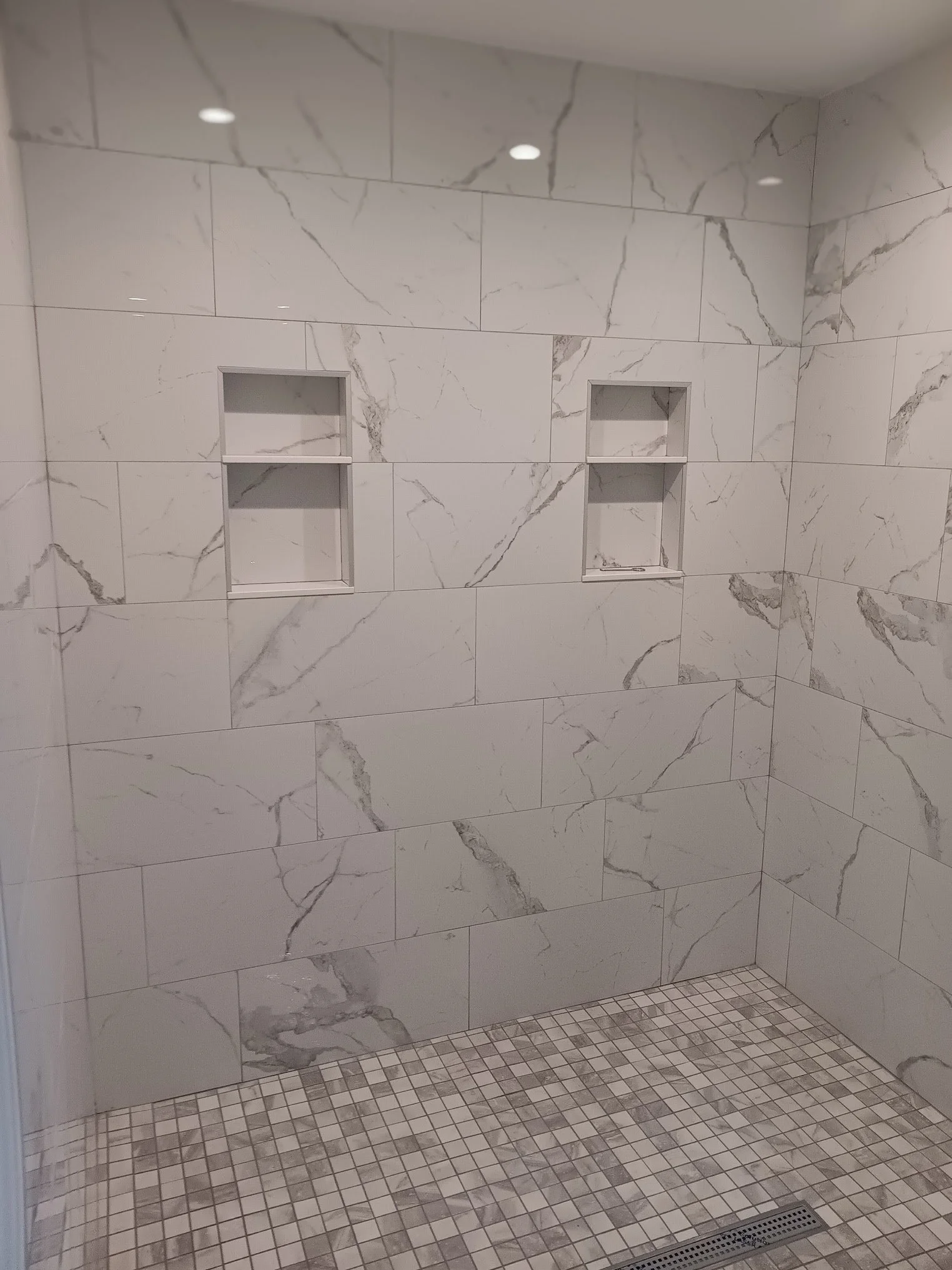 A tiled shower area with marble walls, featuring two recessed shelves, and a floor with small square tiles and a drain.