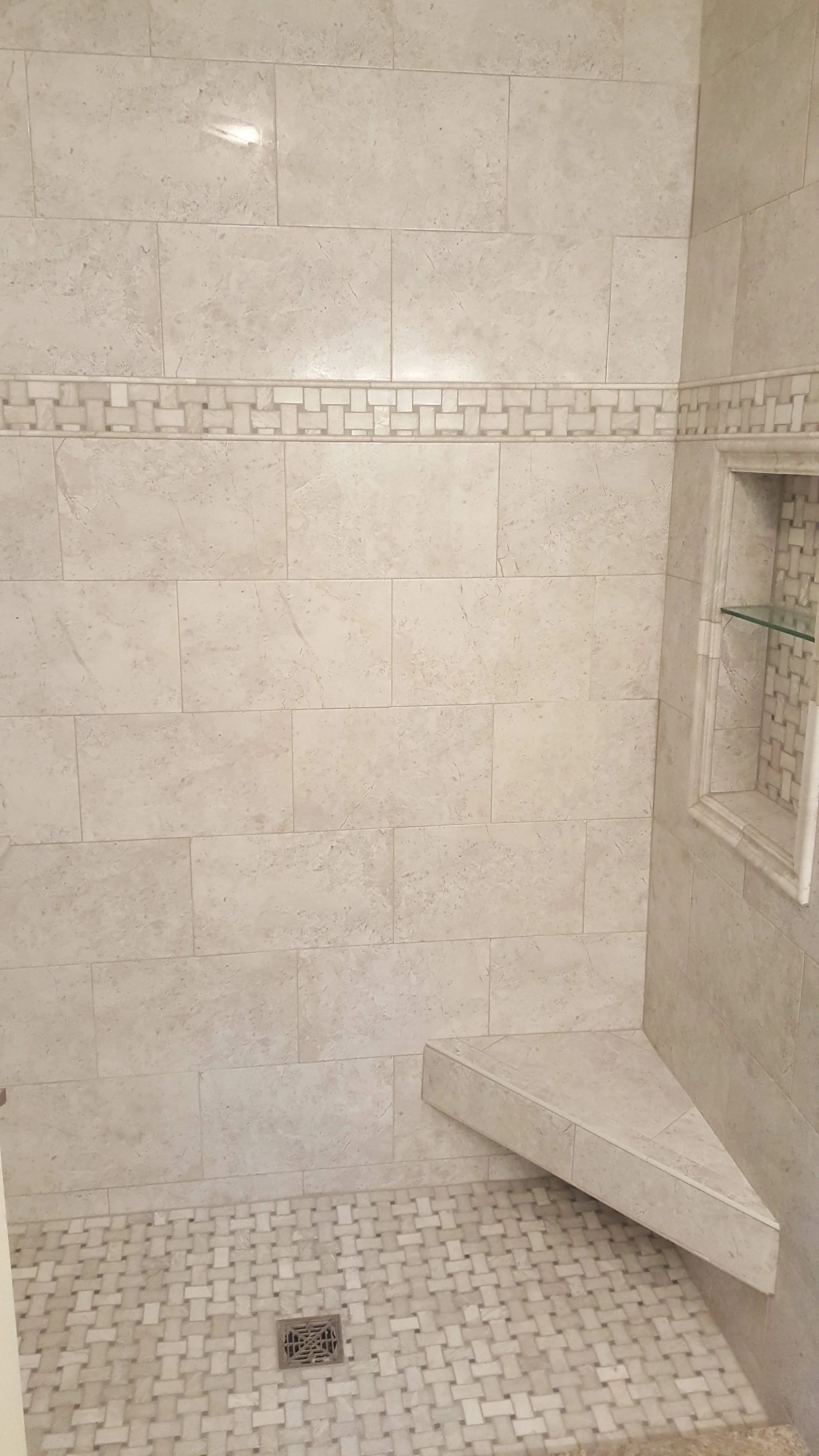 A tiled shower with beige marble tiles on the wall, a decorative tile border, a built-in niche with glass shelves, a corner bench, and a drain on the mosaic tiled floor.