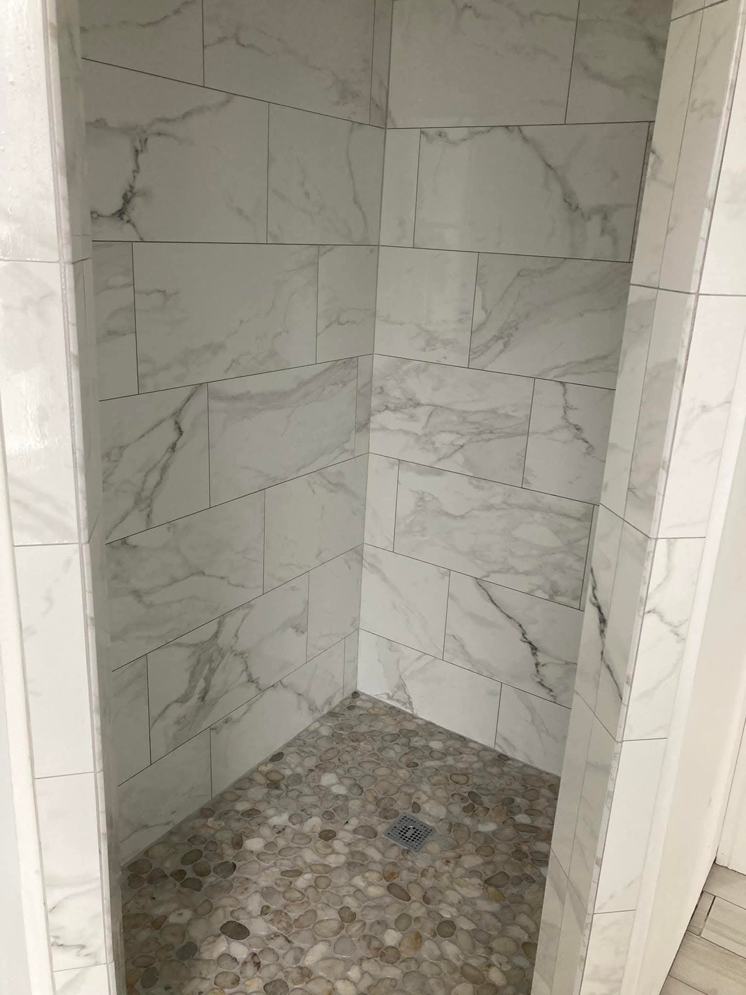 A shower stall with white marble wall tiles featuring gray veining and a pebble stone floor, with a drain on the floor.