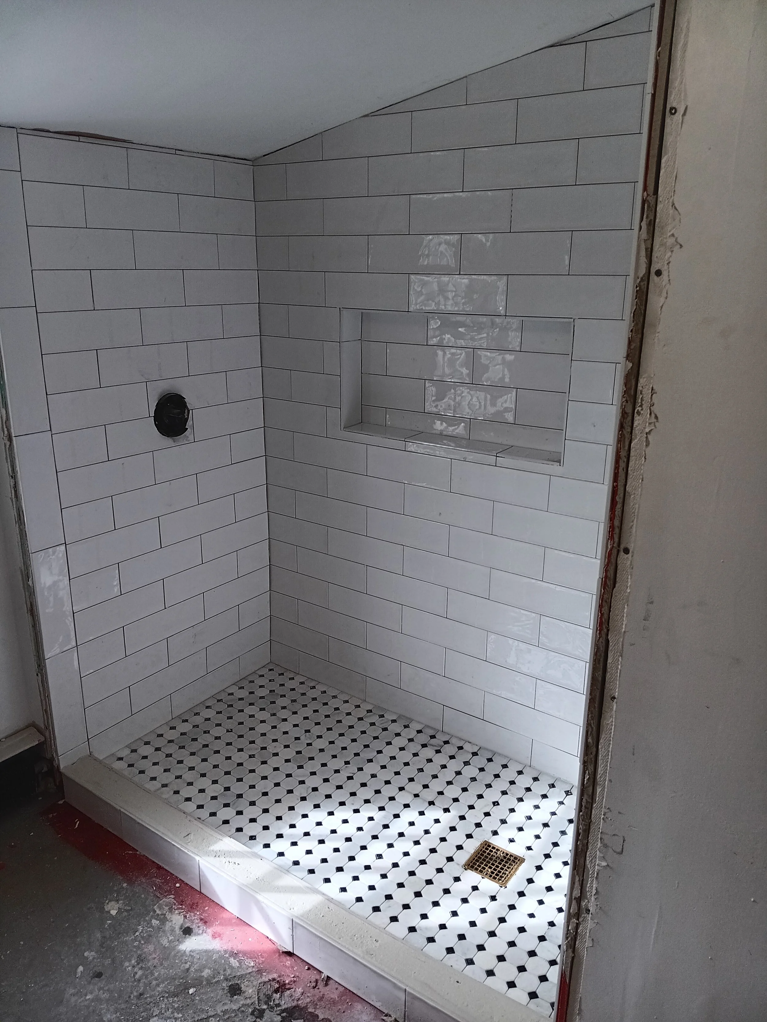 Inside a partially finished bathroom shower with white subway tile on the walls, a small built-in shelf, and black and white honeycomb tile on the floor, with construction materials and unfinished wall edges visible.
