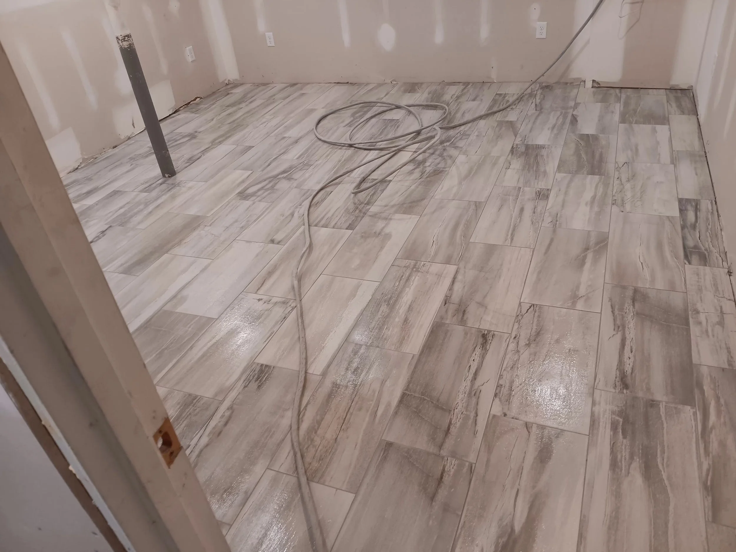 Room with newly installed light-colored wood-look tile flooring, unfinished drywall on walls with electrical outlets, and construction tools including a metal pipe, a coil of electrical wire, and a power cord.