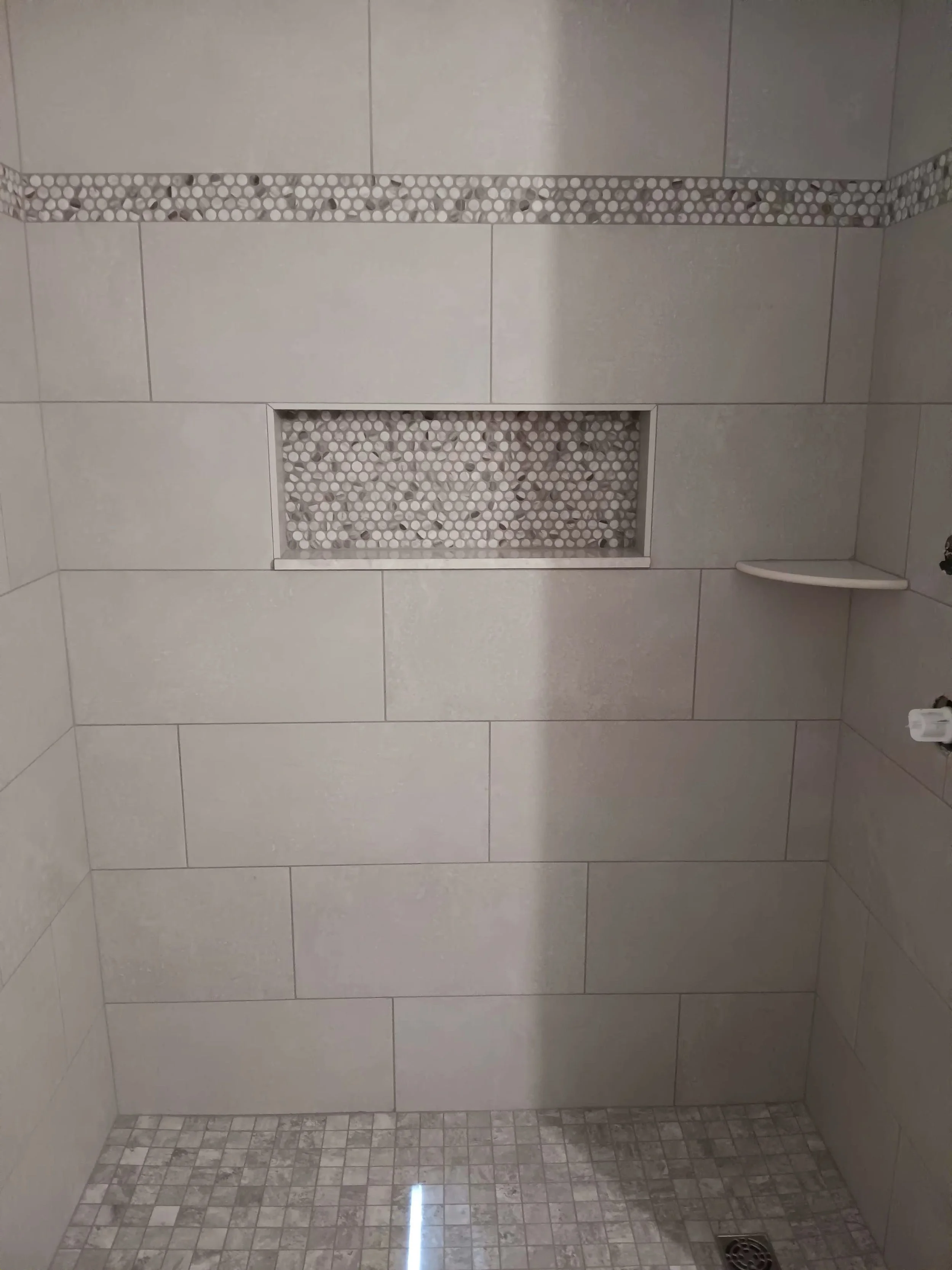 A tiled shower wall with a mosaic tile border, a rectangular niche with pebble tiles, a small shelf in the corner, and a floor with smaller square tiles