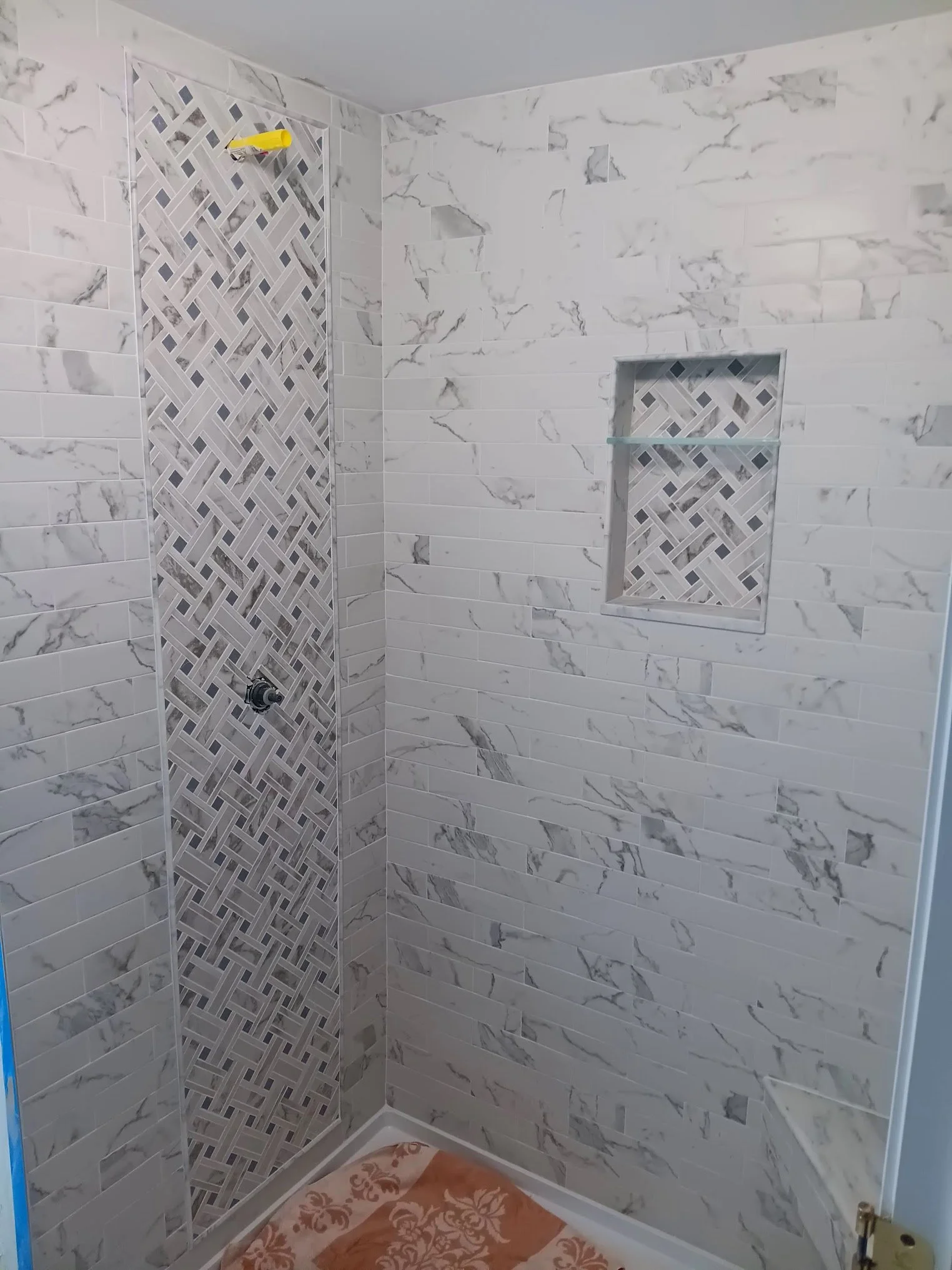 A walk-in shower with white marble-like wall tiles and a vertical accent strip of intricate gray and white mosaic tiles. There is a recessed niche with matching mosaic tiles and a glass shelf. A yellow scraper tool is attached to the wall. The corner