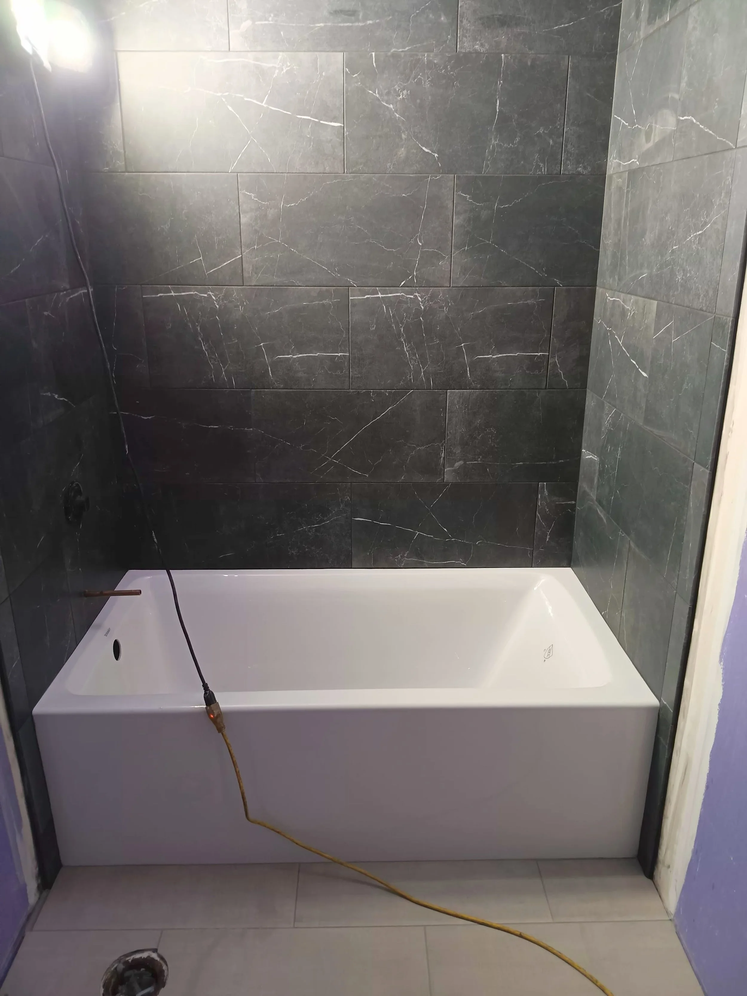 Bathroom with black marble tile walls and a white bathtub, with a yellow electrical cord hanging over the tub.
