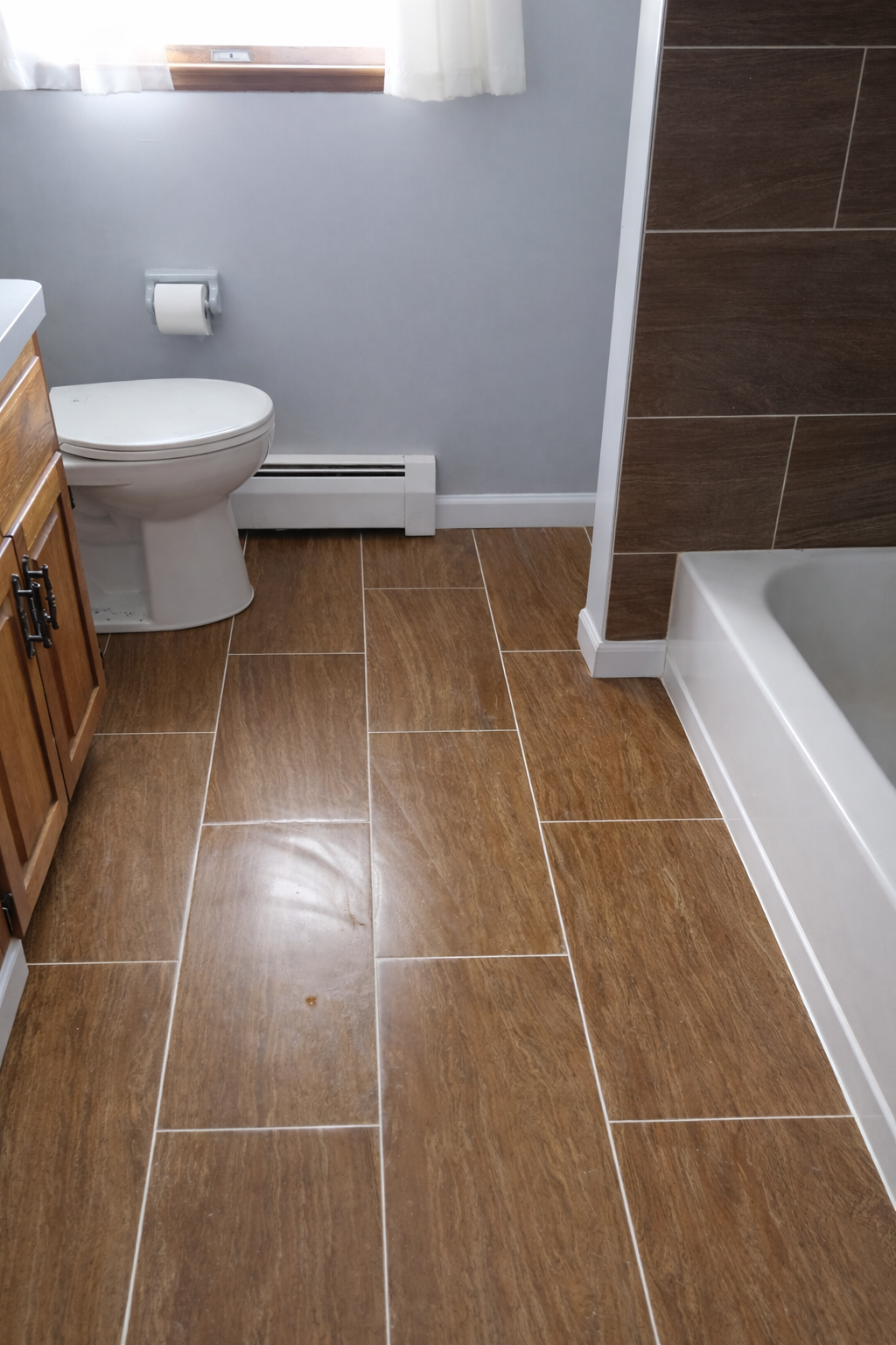 Bathroom with tan floor tiles, a white toilet, a wooden cabinet, a baseboard heater, a window with white curtains, and a shower area with brown tiles.
