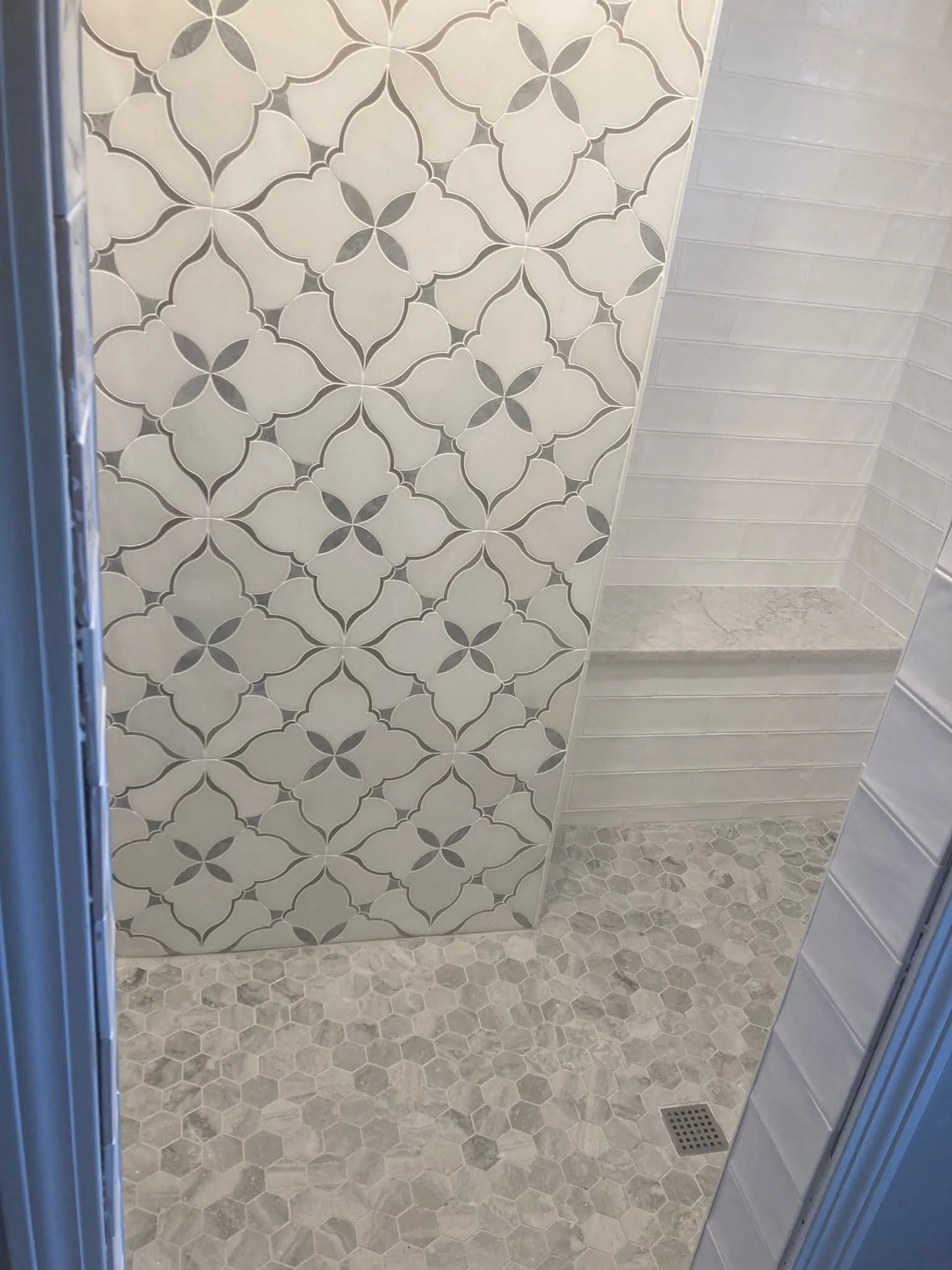 A bathroom shower area with a decorative patterned tile wall and a sink with a light gray tiled wall and small hexagonal floor tiles.
